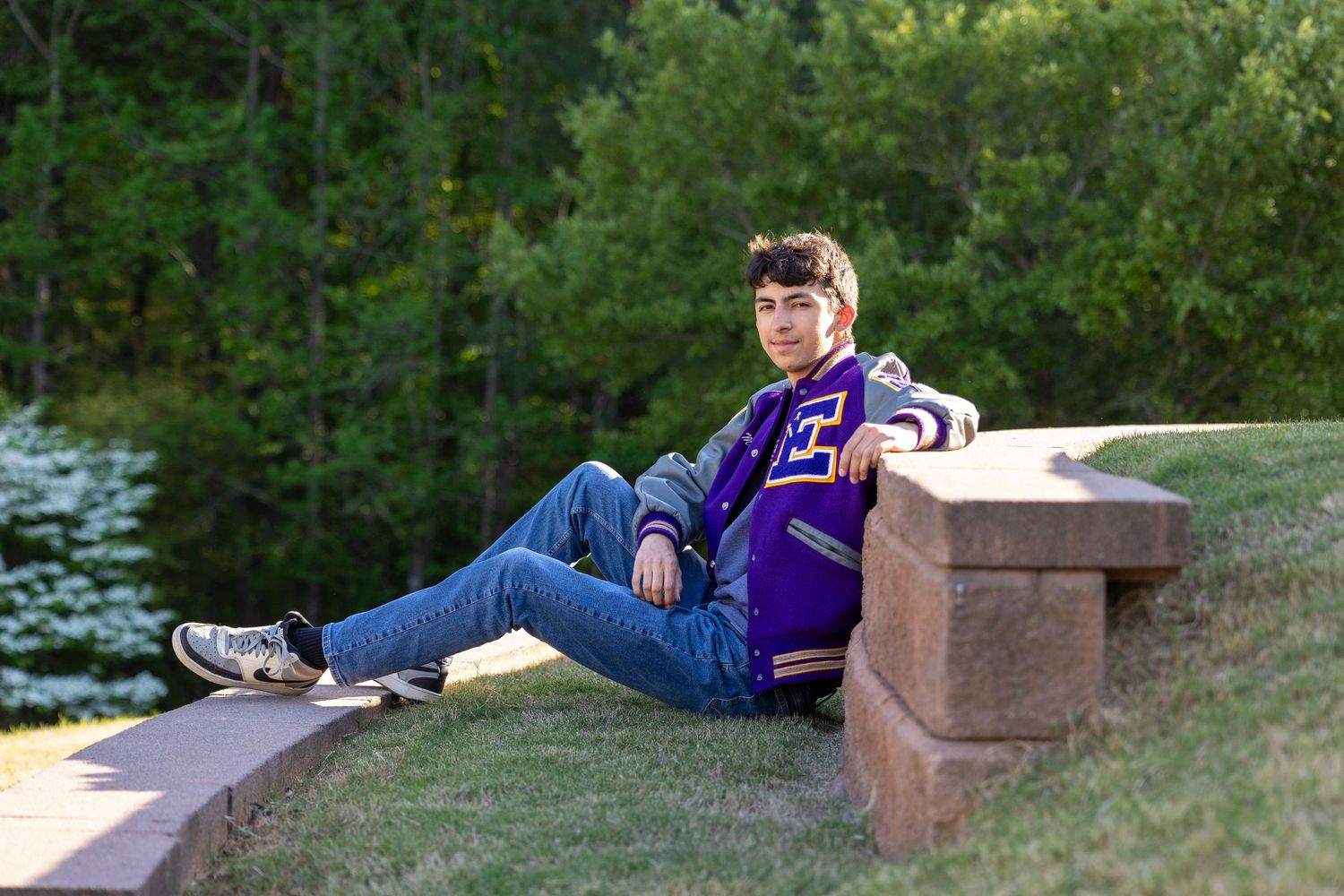 Teen in purple letterman jacket poses on stone wall with lush green trees in background for senior portrait.