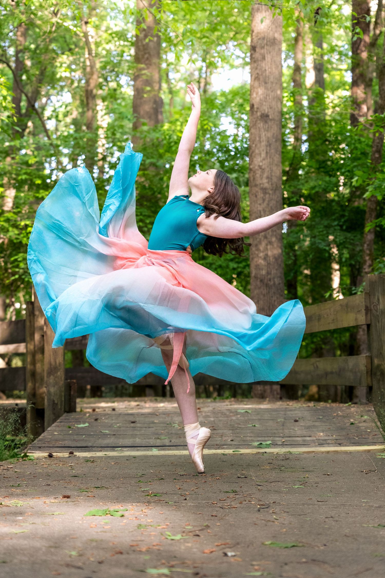 A ballerina in a flowing pink and blue skirt dances on pointe in a sunlit forest path.
