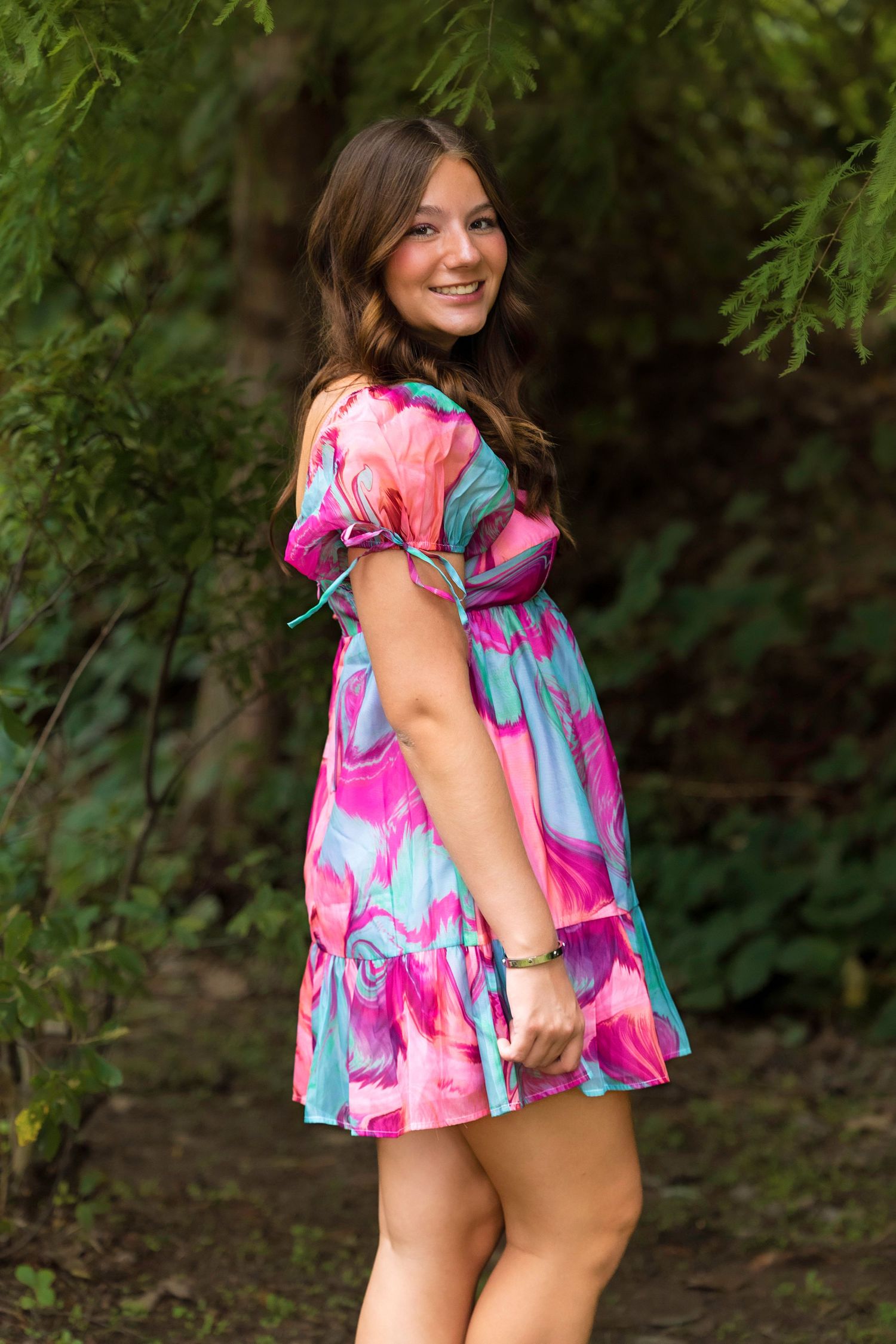 Young woman smiling in a pink and teal floral mini dress, posing outdoors among lush green trees.