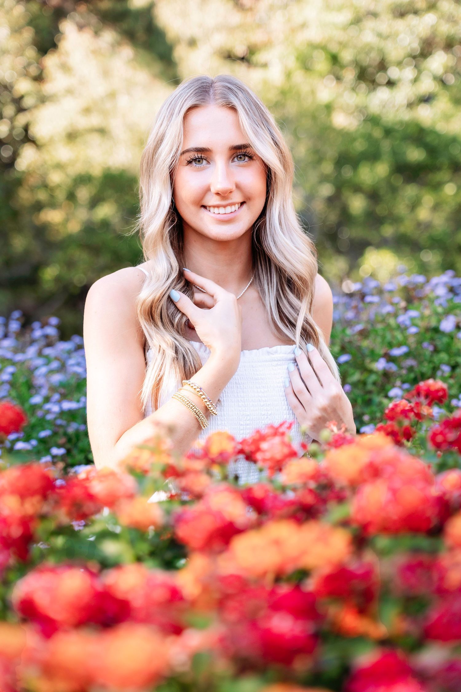 A woman poses in a white top surrounded by vibrant red and purple flowers in a sunny garden setting.