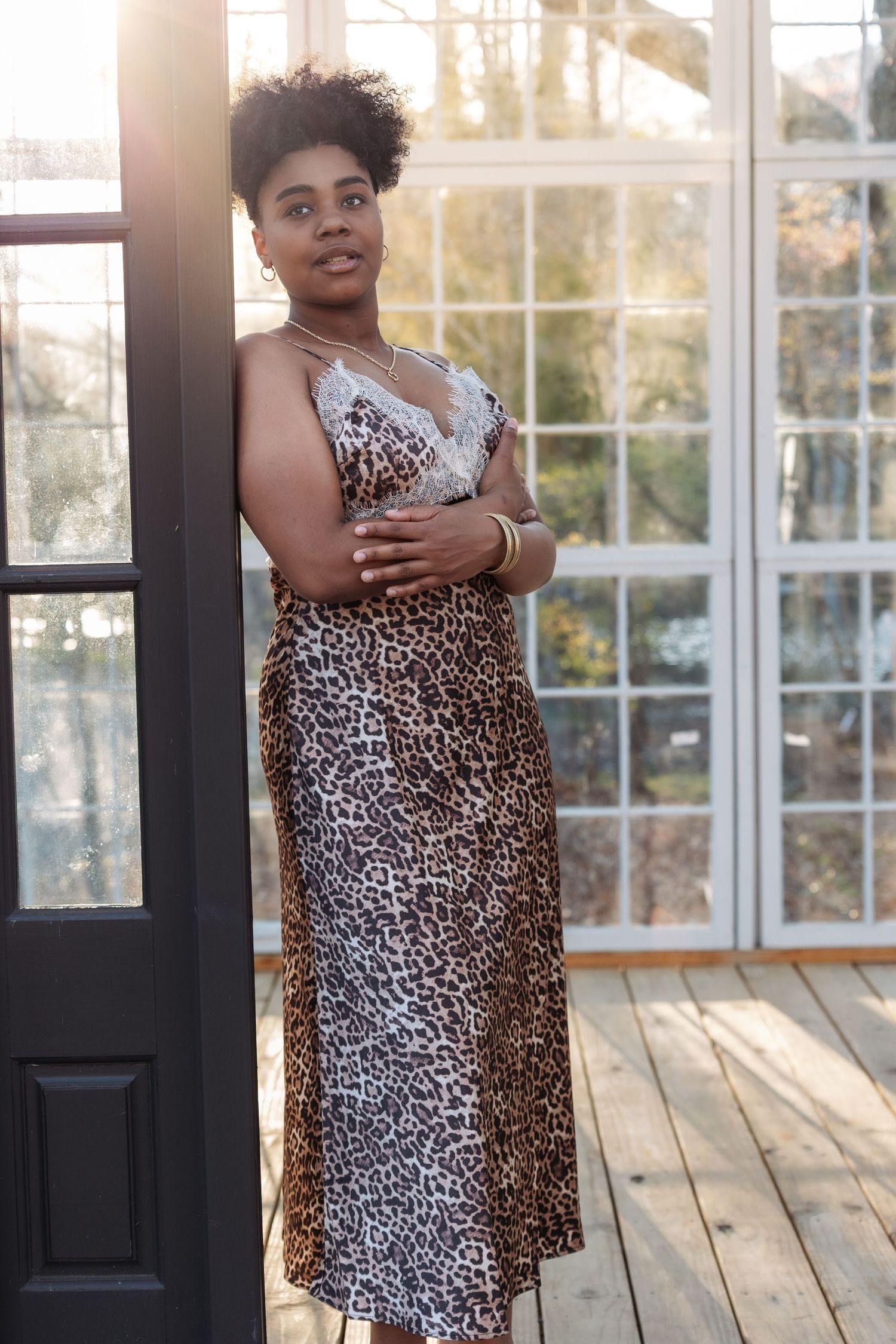 Woman in leopard print midi dress posing by black door in sunlit glass room.