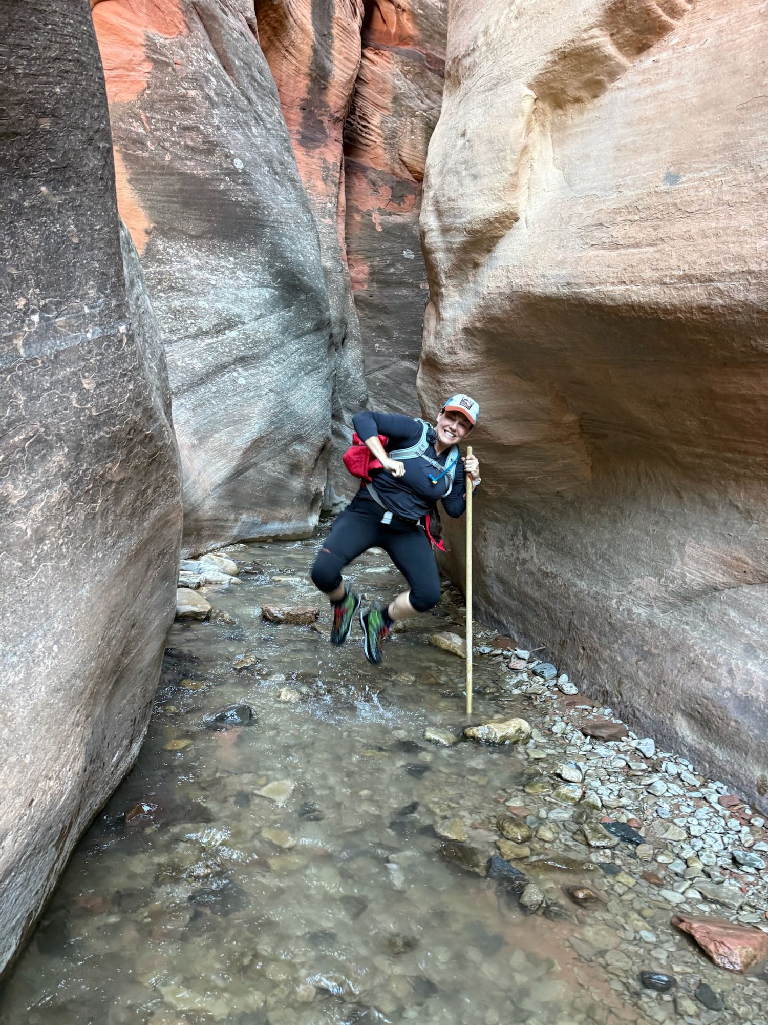 Hiker with walking stick navigating a narrow slot canyon with shallow water and red sandstone walls.