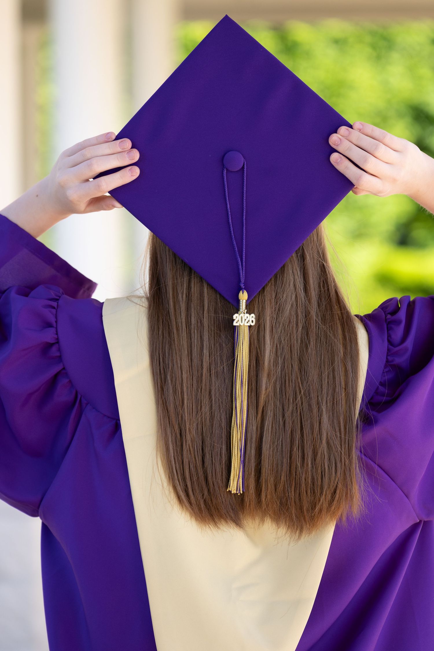 Back view of graduate in purple cap and gown holding mortarboard with gold 2025 tassel outdoors.