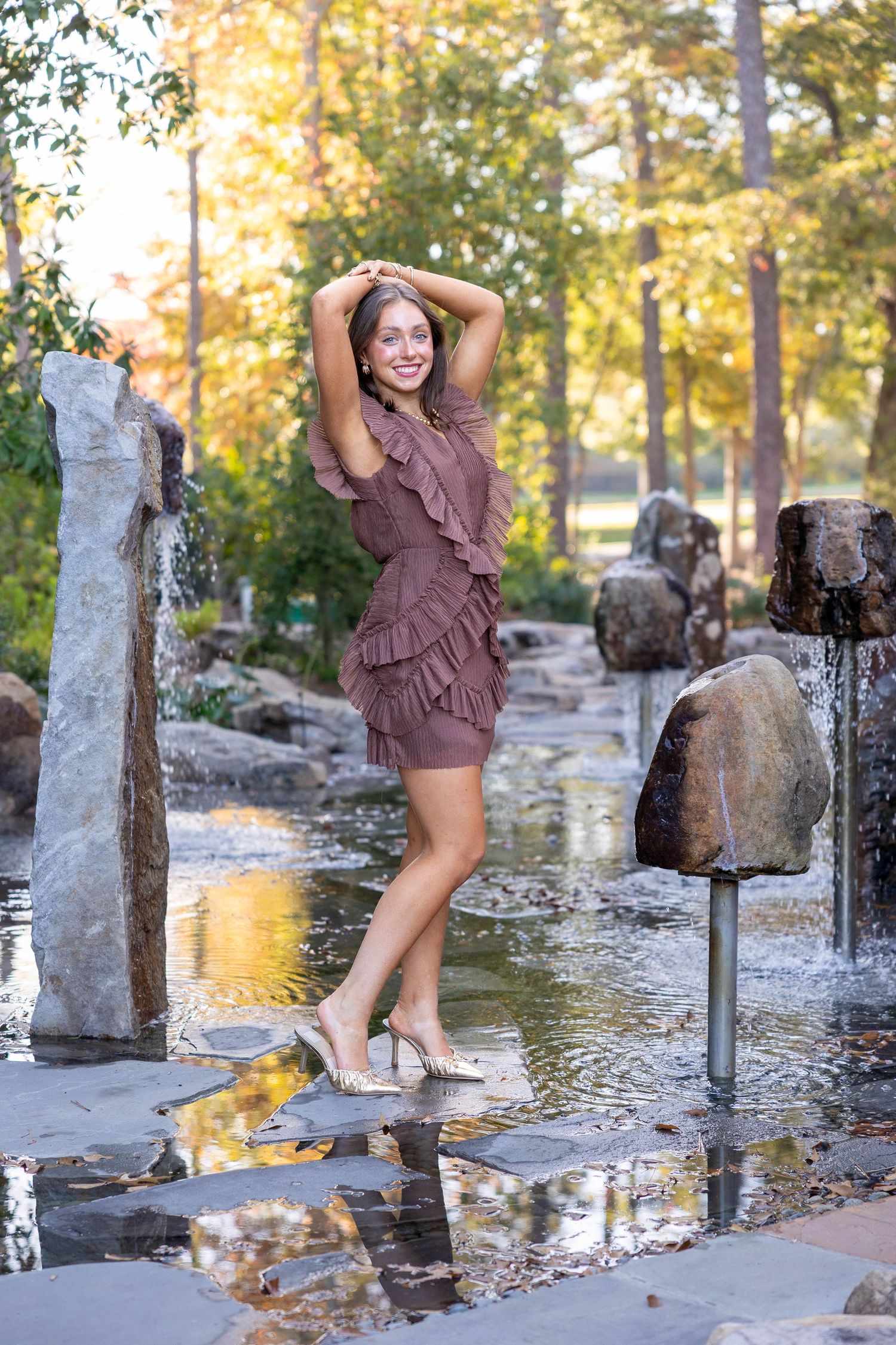 A person in a brown dress poses playfully near a water feature in a park during golden hour lighting.