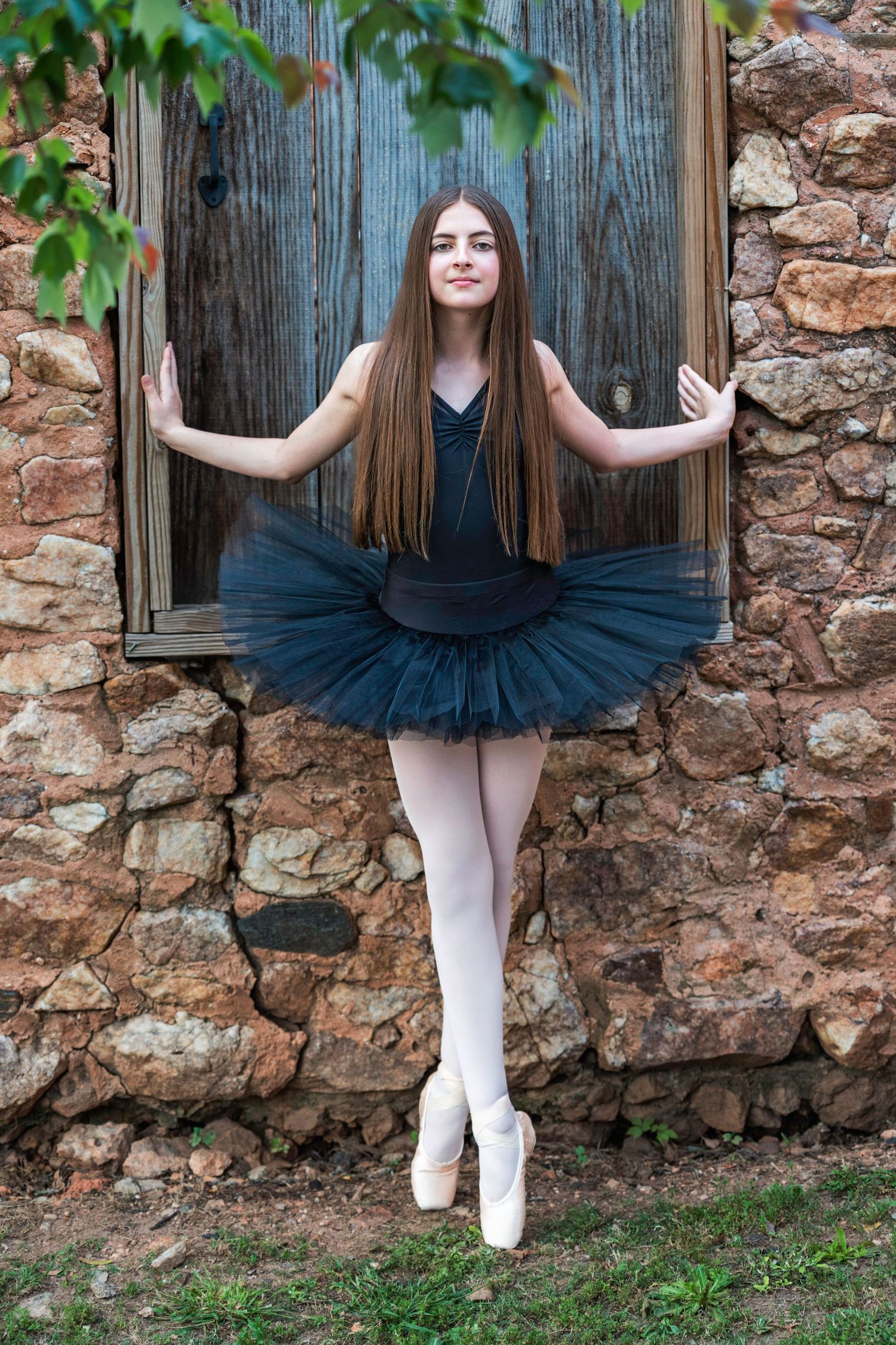 A ballet dancer in a black tutu and pointe shoes poses gracefully against a rustic wooden door and stone wall.