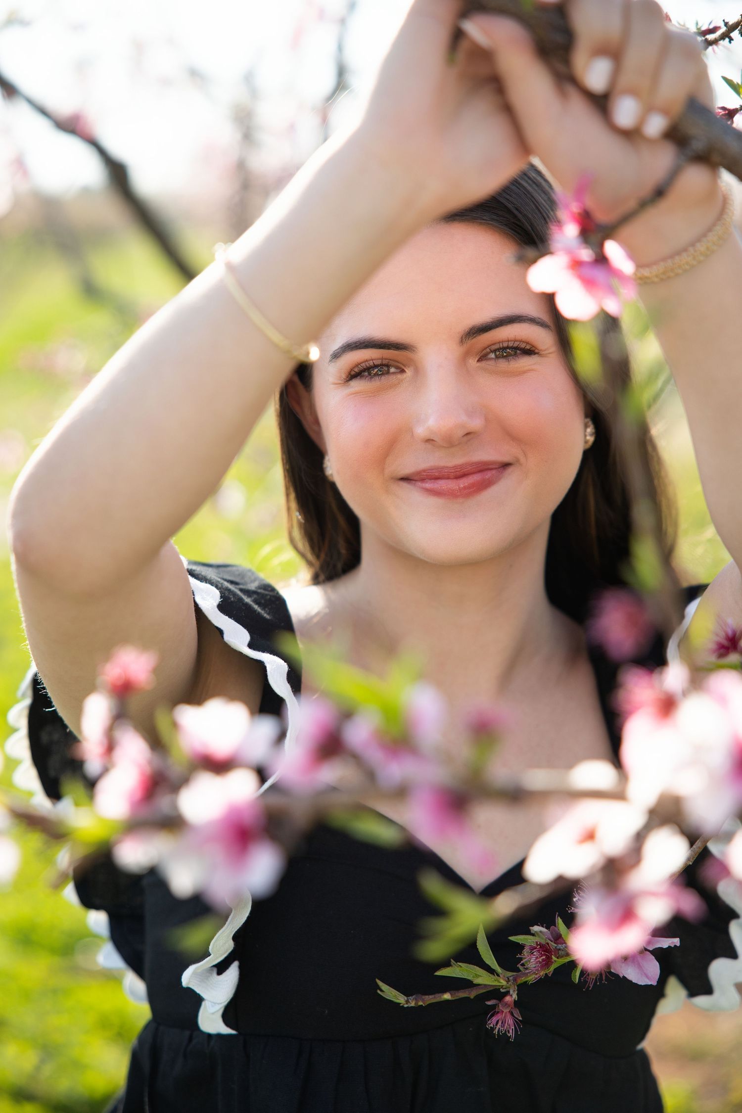 A person in black poses among delicate pink cherry blossoms on a bright spring day.