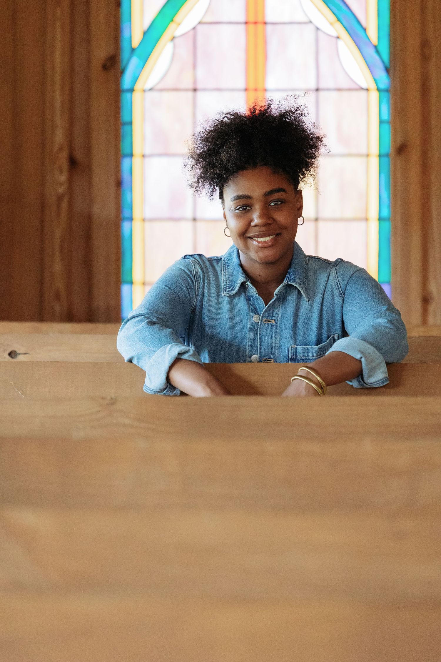 Smiling young Black woman in denim shirt leaning on wooden pew in front of colorful stained glass window.