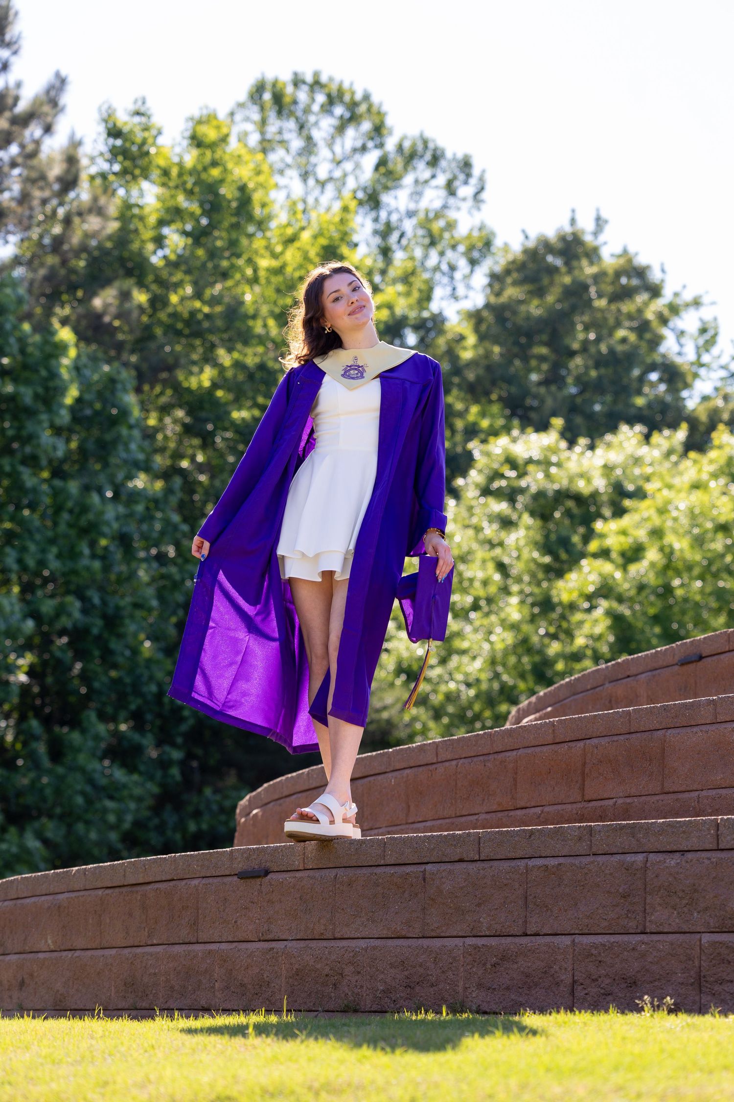 Young woman in purple graduation gown and cap stands on stone steps outdoors with green trees behind her.