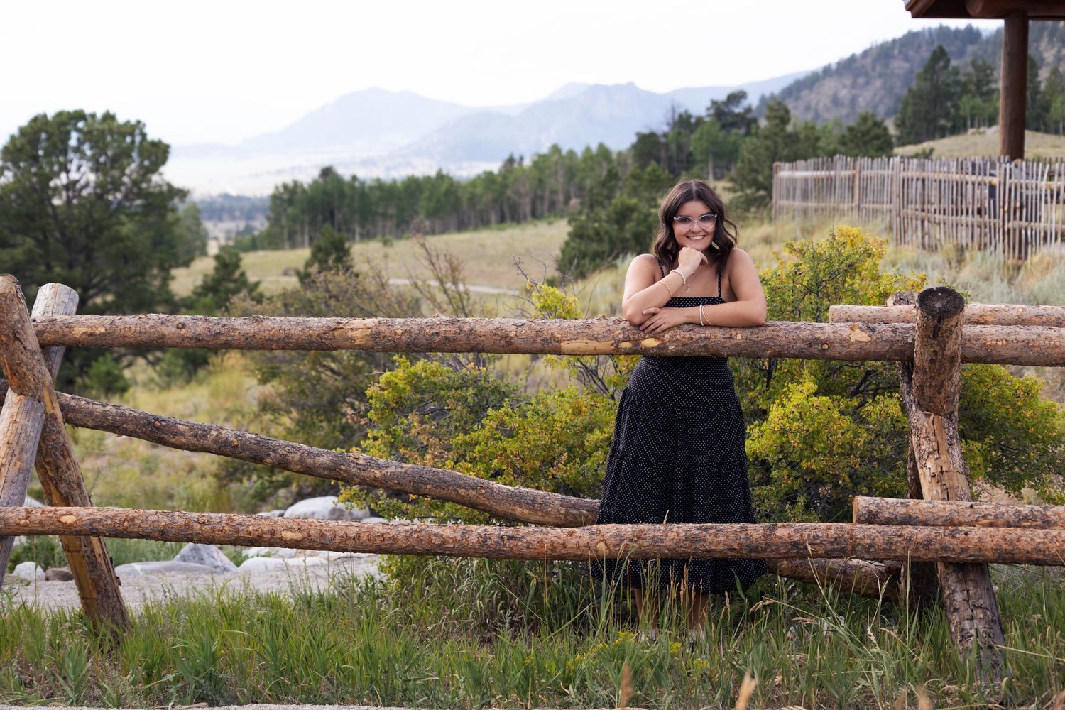 Person posing near rustic wooden fence with mountain landscape in background.
