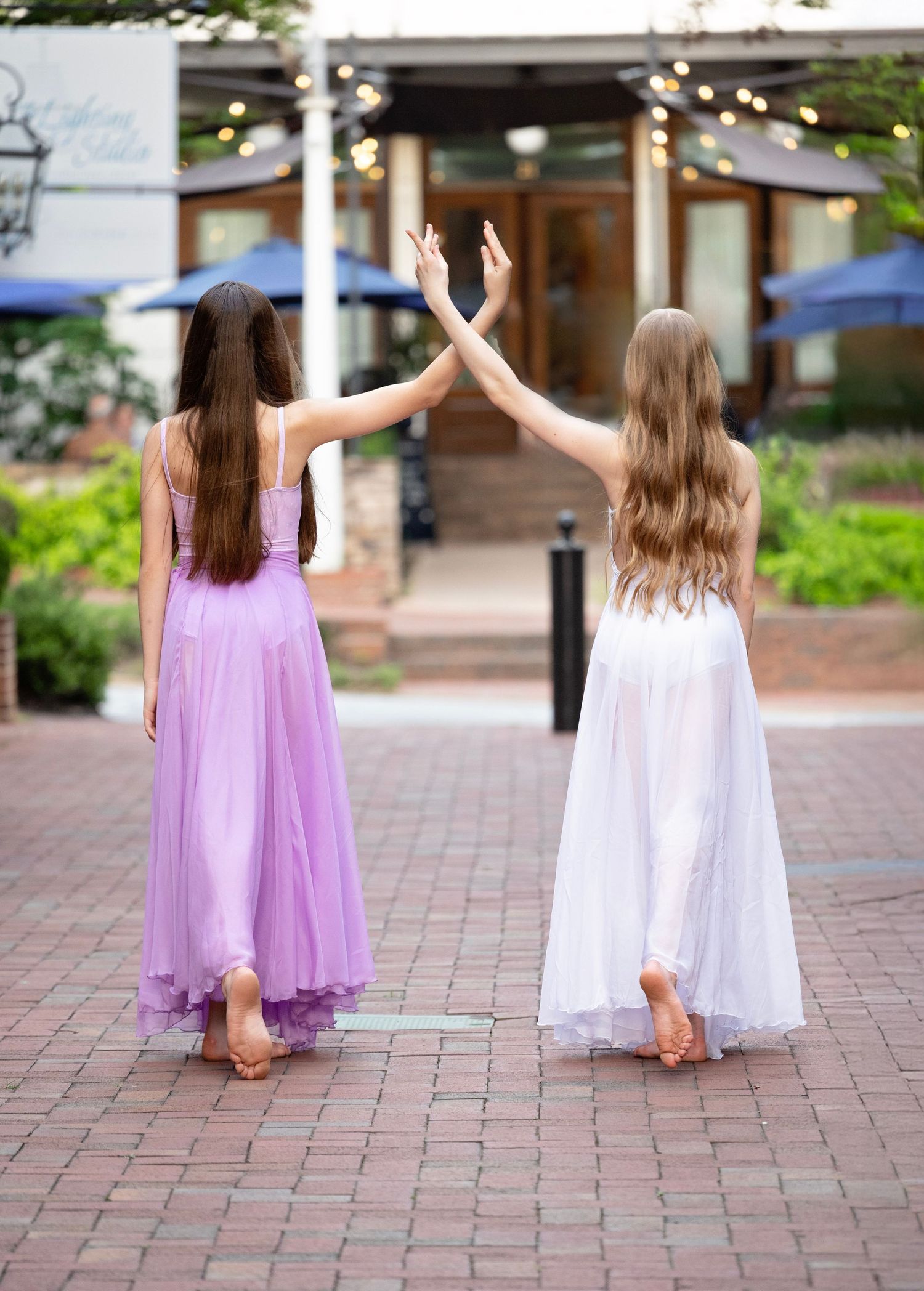 Two children in flowing dresses stand back-to-back with raised arms on a brick courtyard.