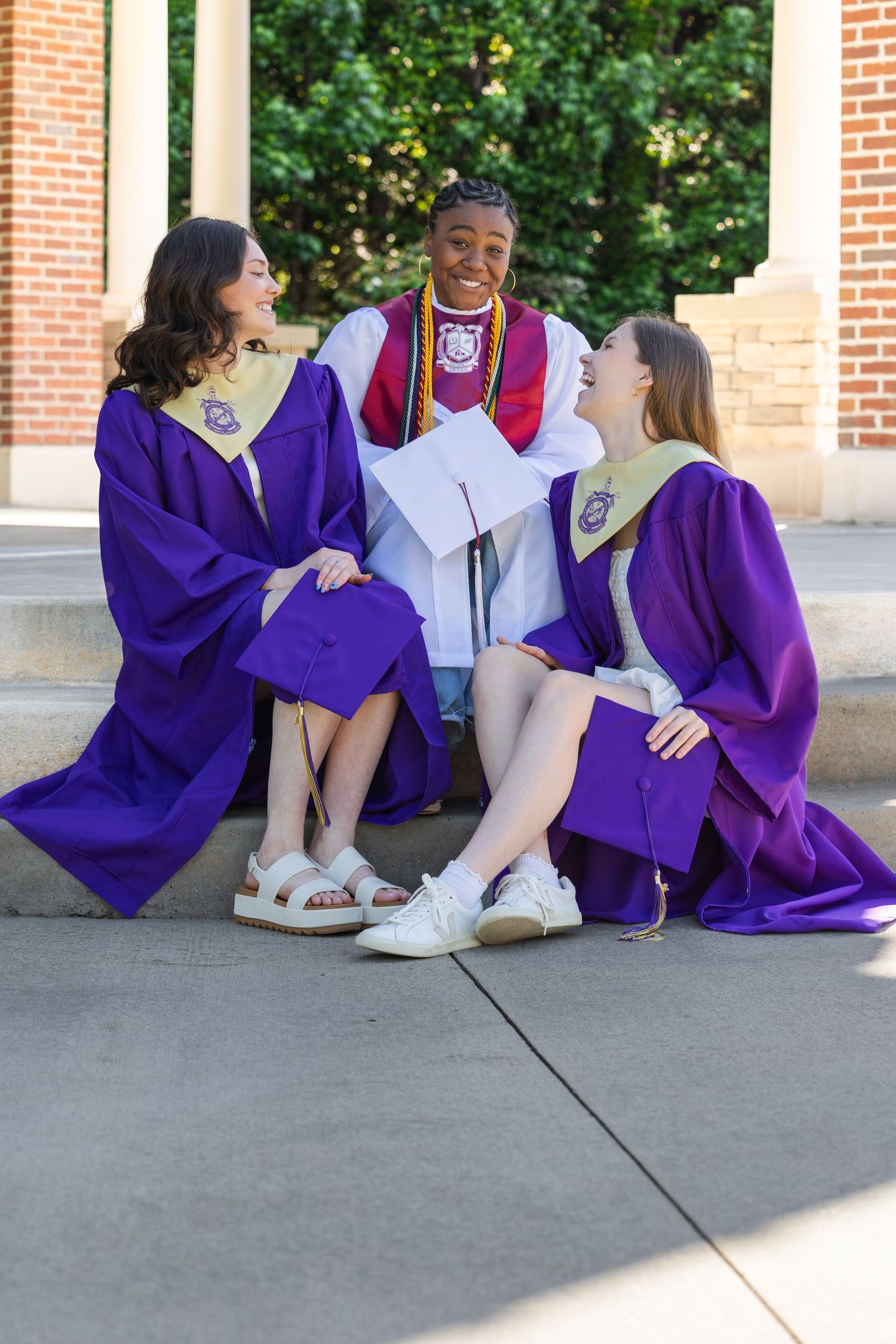 Three graduates in purple robes sit together on steps, smiling and celebrating their graduation day.