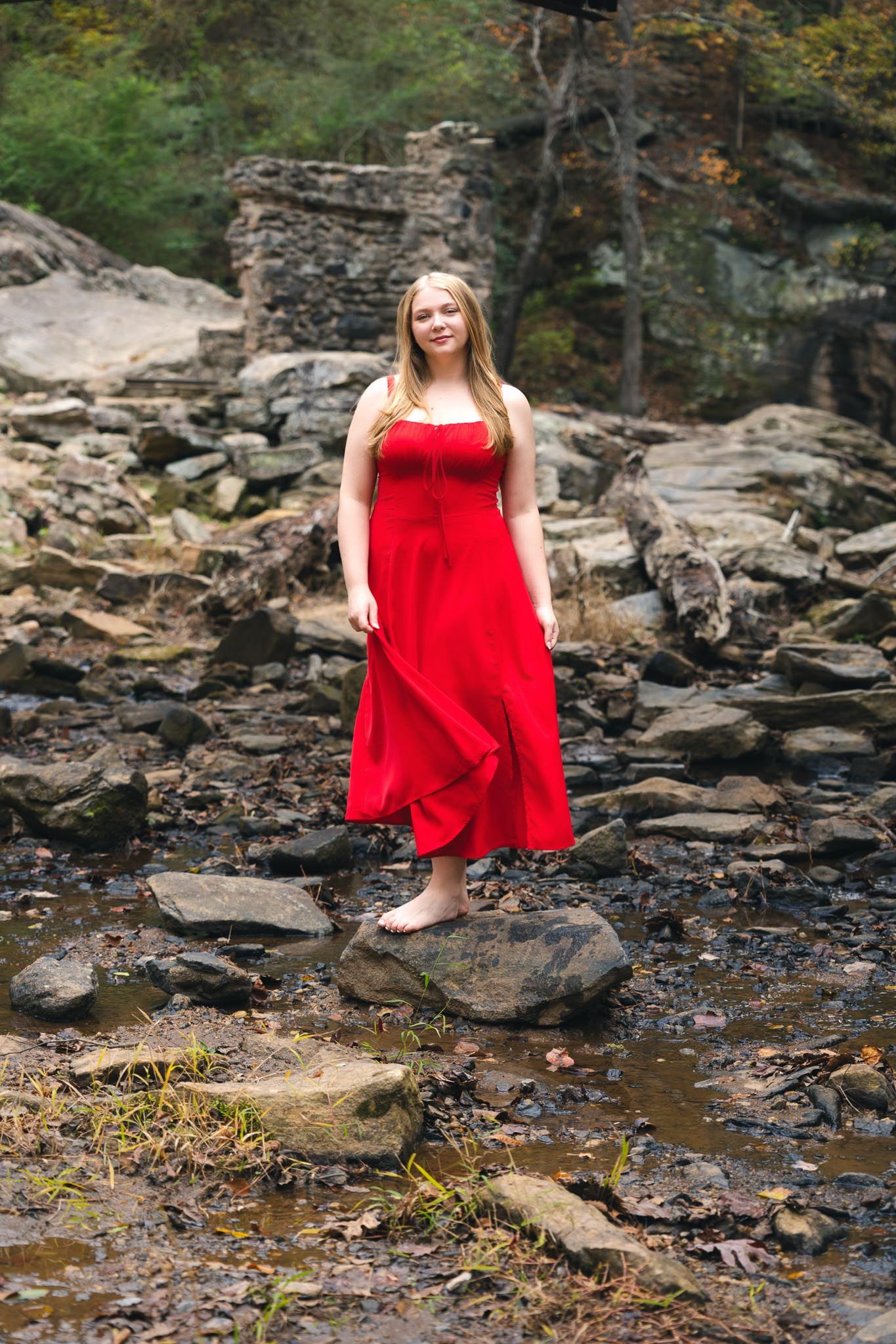 A person in a flowing red dress poses gracefully on rocky terrain near a stream in a wooded setting.