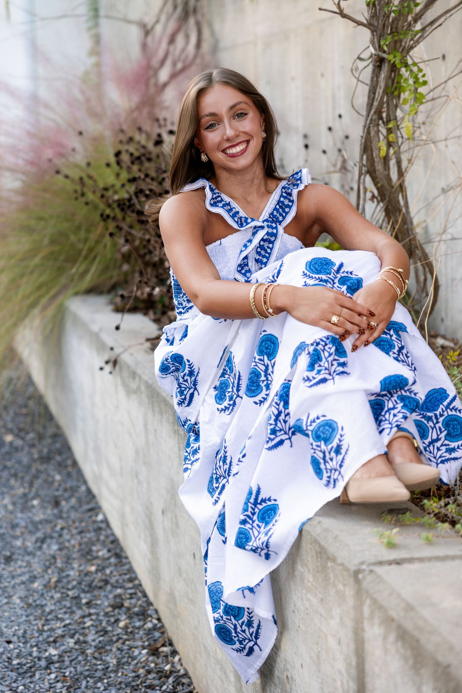 A summer dress in white and blue floral print is worn while sitting on concrete steps near decorative grasses.