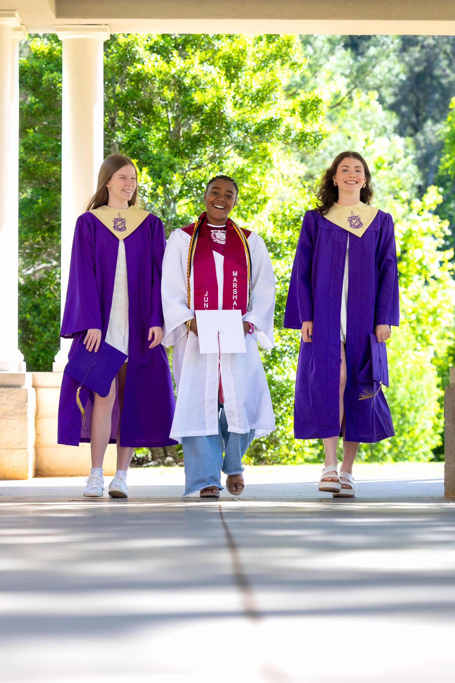 Three graduates in purple and white robes walk together outdoors, smiling during a graduation ceremony.