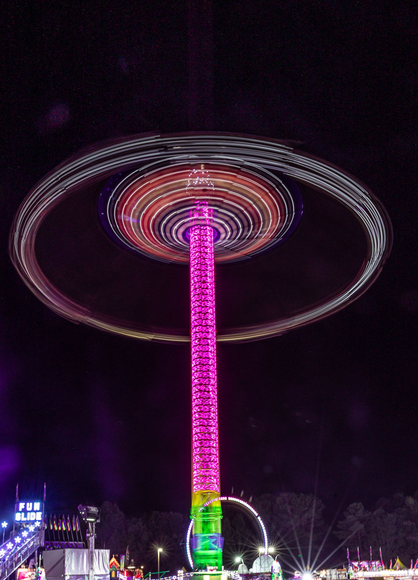Long exposure night photo of spinning carnival ride creating circular pink light trails.