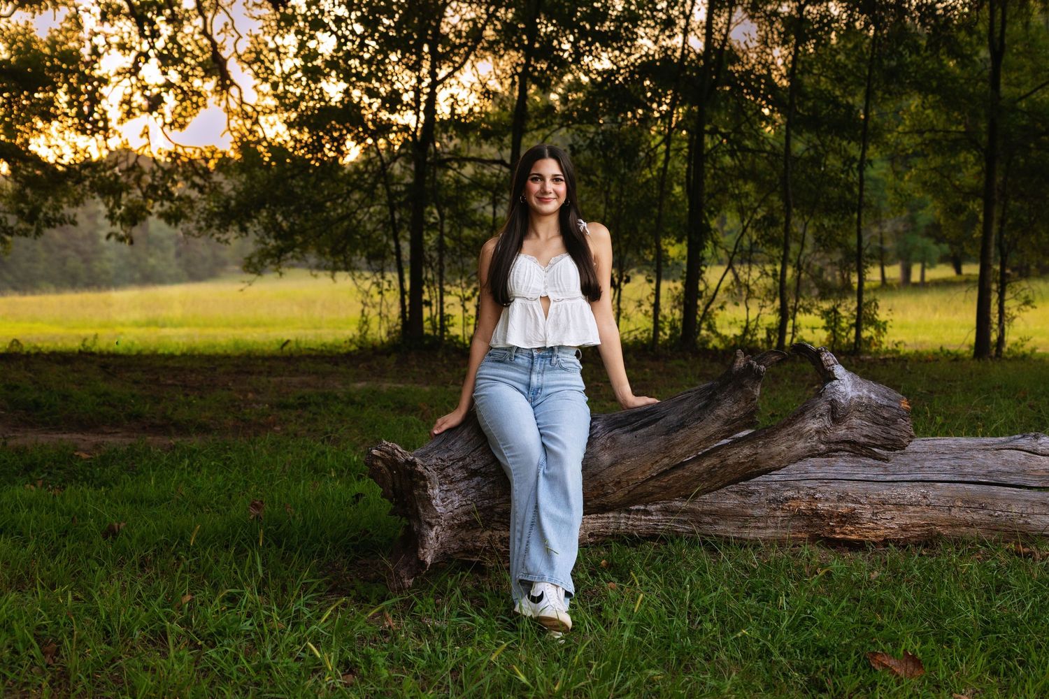 Young woman in white top and jeans leaning on a fallen log in a lush green park at golden hour.