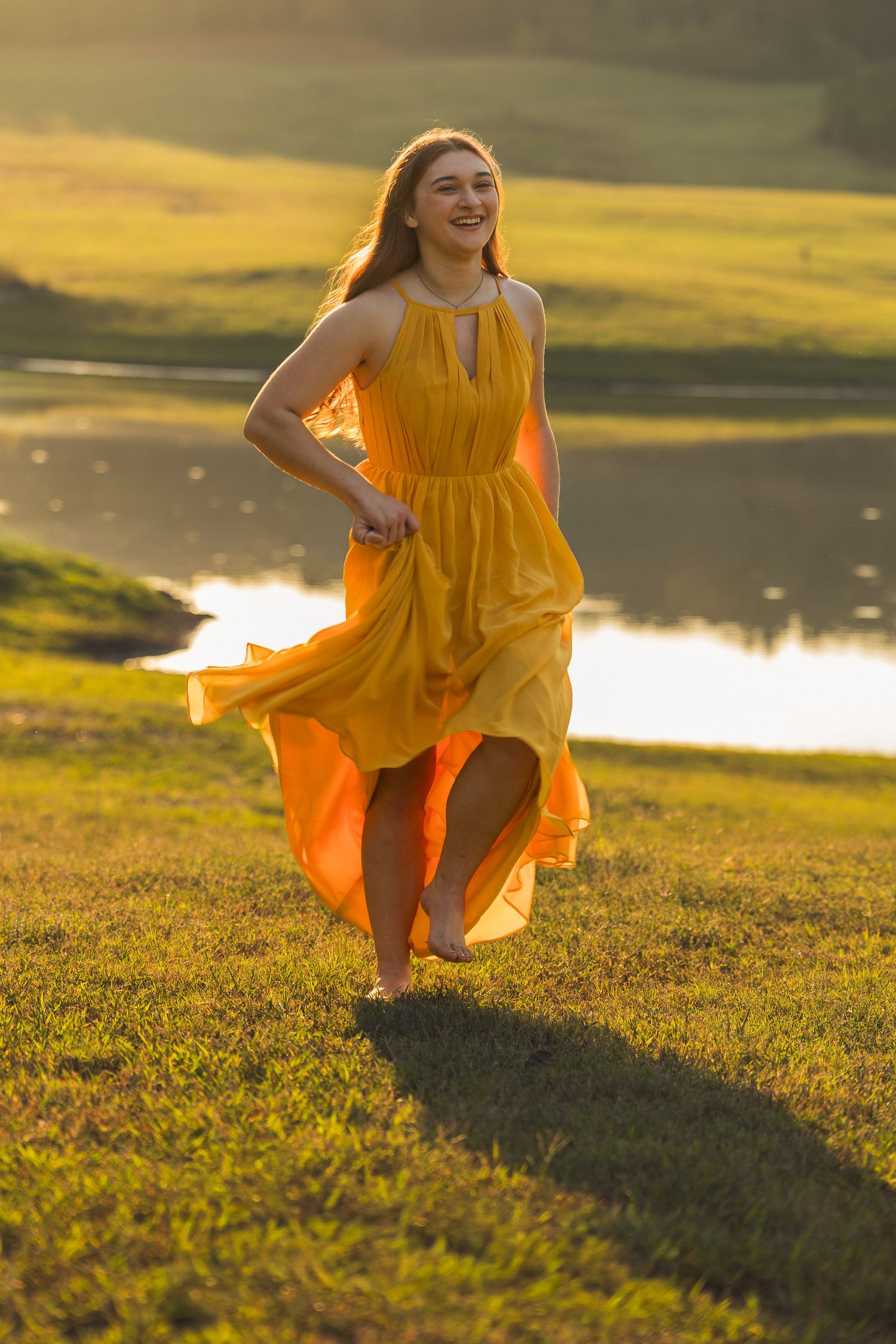 Young woman in flowing yellow dress dancing barefoot on green grass near a sunlit lake at golden hour.