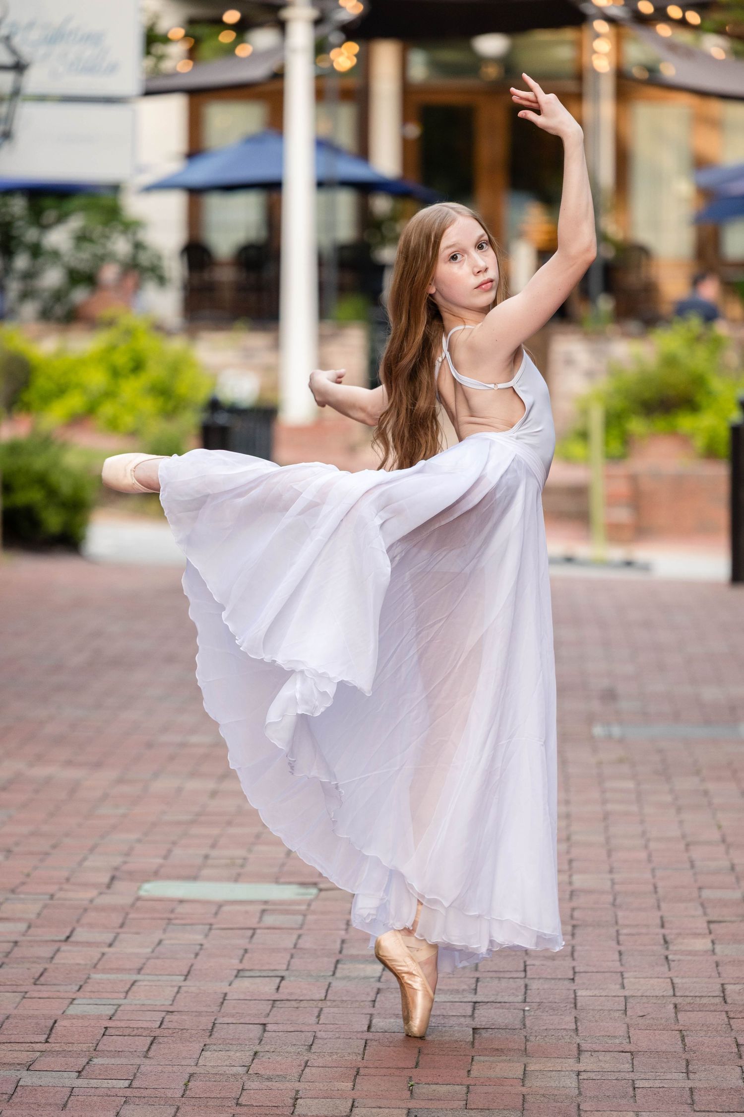 A dancer in a flowing white dress performs graceful ballet movements on a brick courtyard.