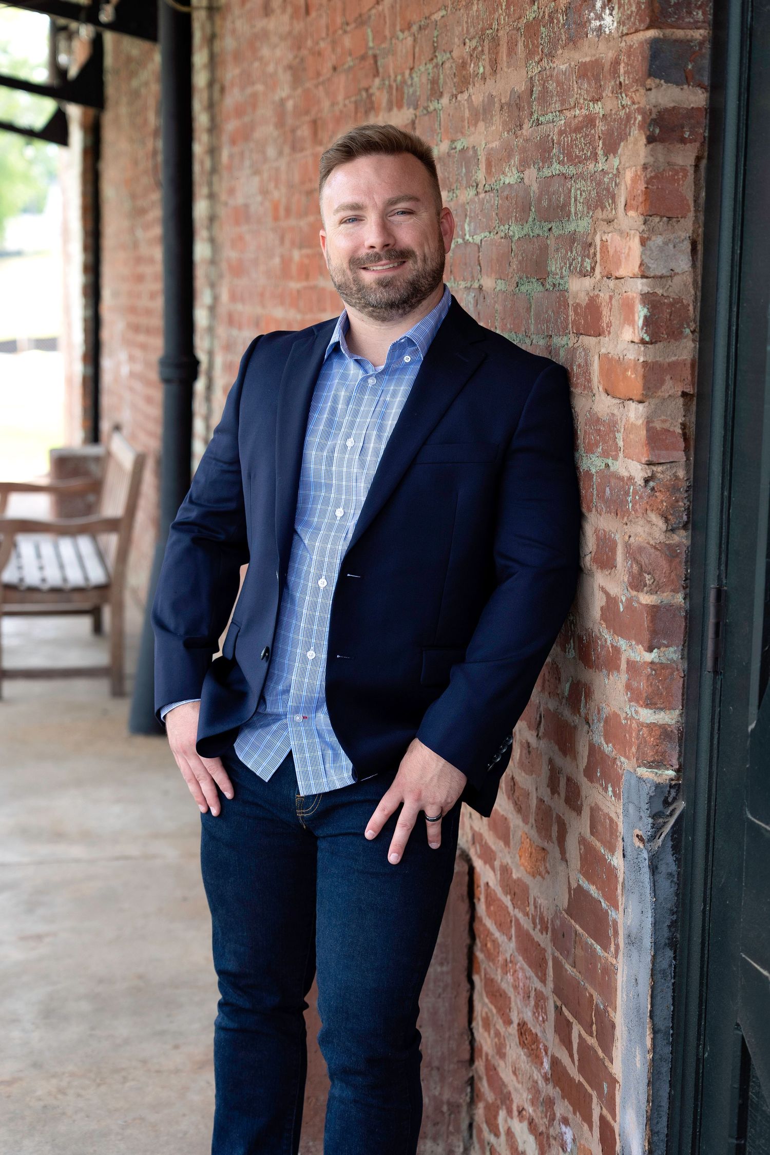 Professional business portrait of someone in a navy suit and blue checkered shirt standing against a brick wall background.