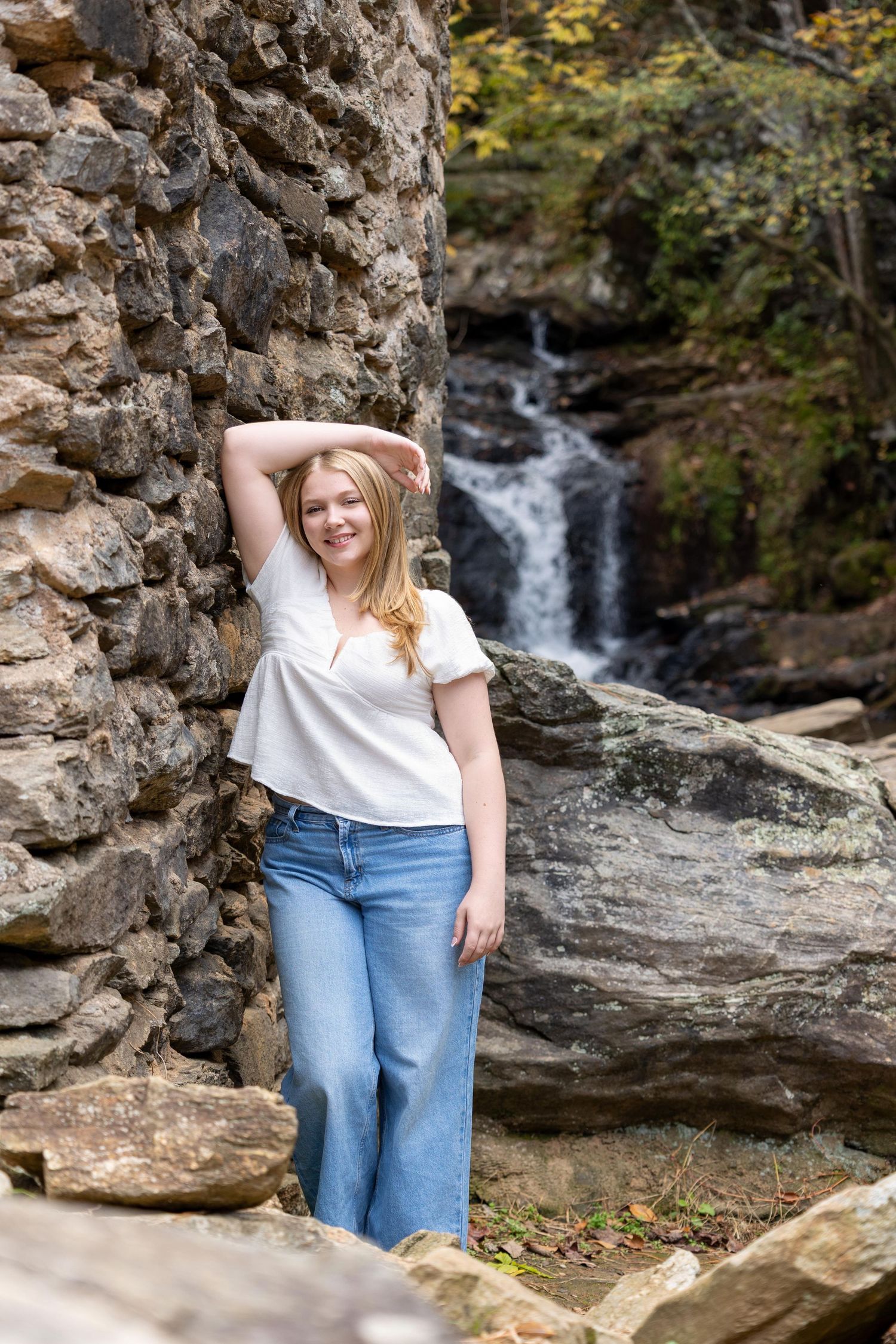 A person in jeans and white shirt poses near a cascading waterfall surrounded by rocky terrain and lush foliage.