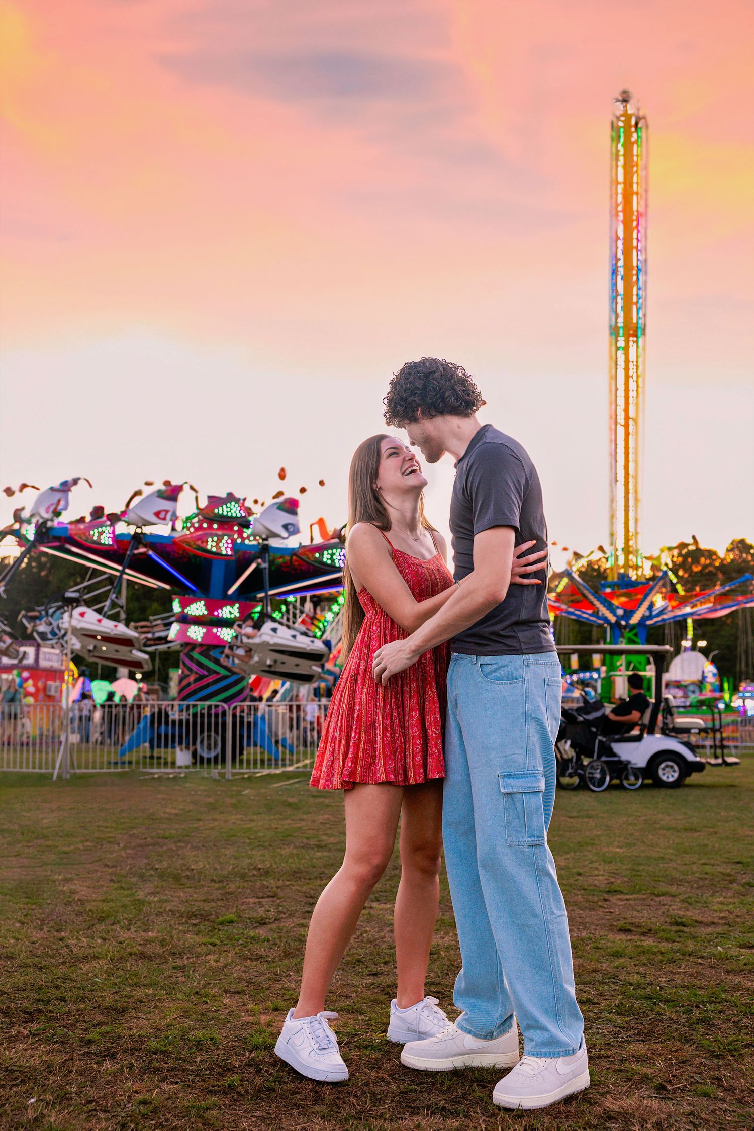 A couple shares a romantic moment at sunset with carnival rides in the background.