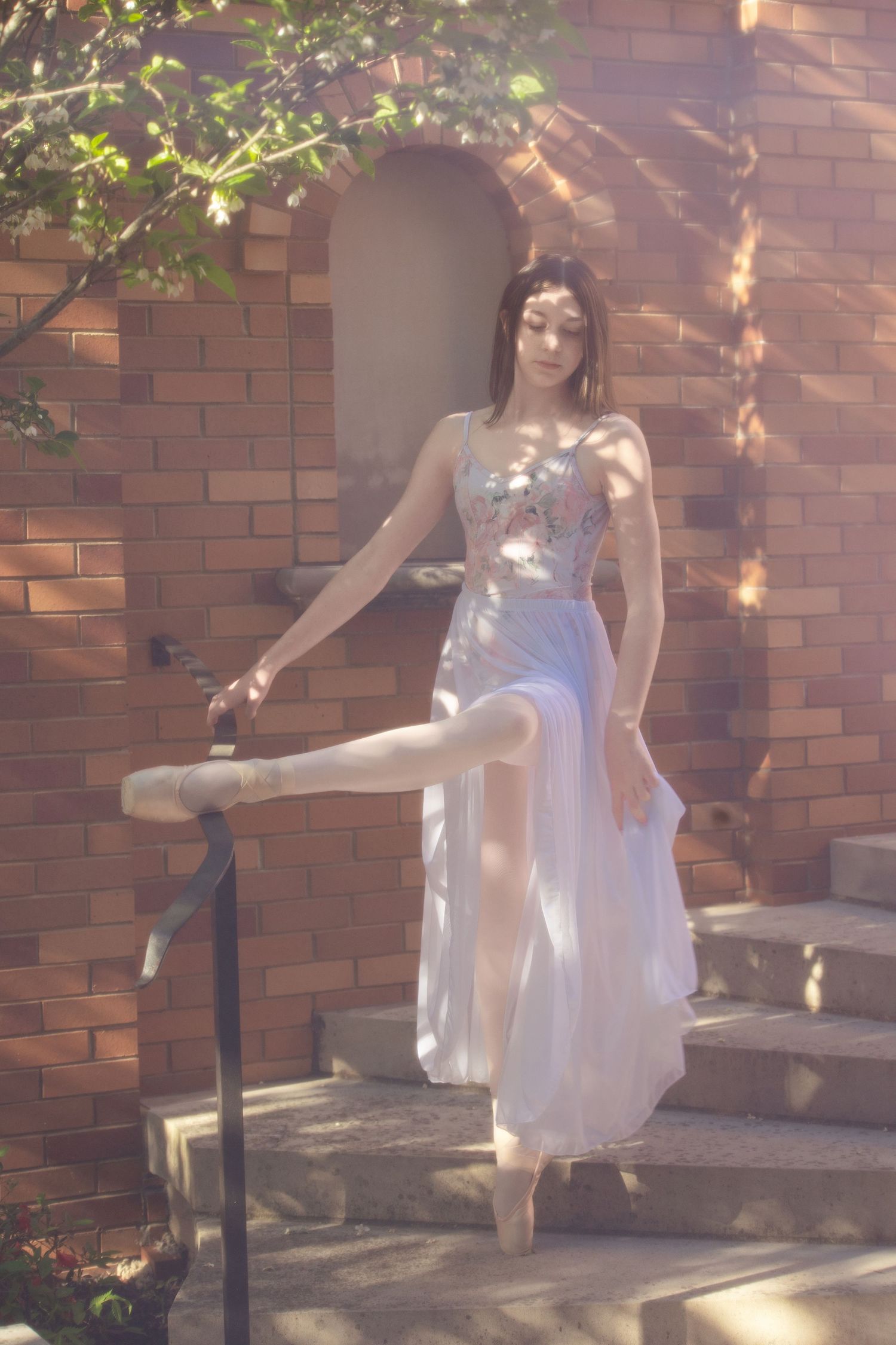 Dancer in flowing white dress poses gracefully on brick steps with dappled sunlight.
