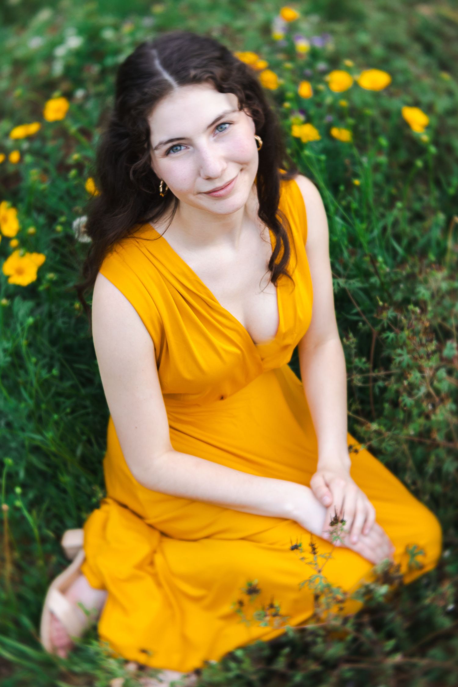 Young woman in mustard yellow dress sitting among wildflowers and green grass, smiling softly at camera.