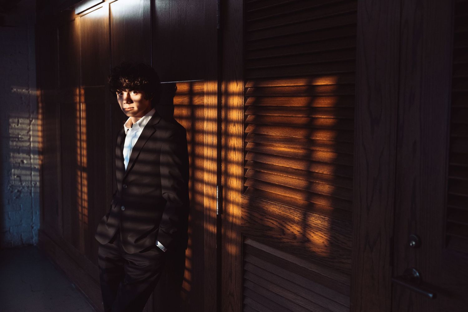 A man in a dark suit stands against a wall with dramatic light and shadow stripes from window blinds.