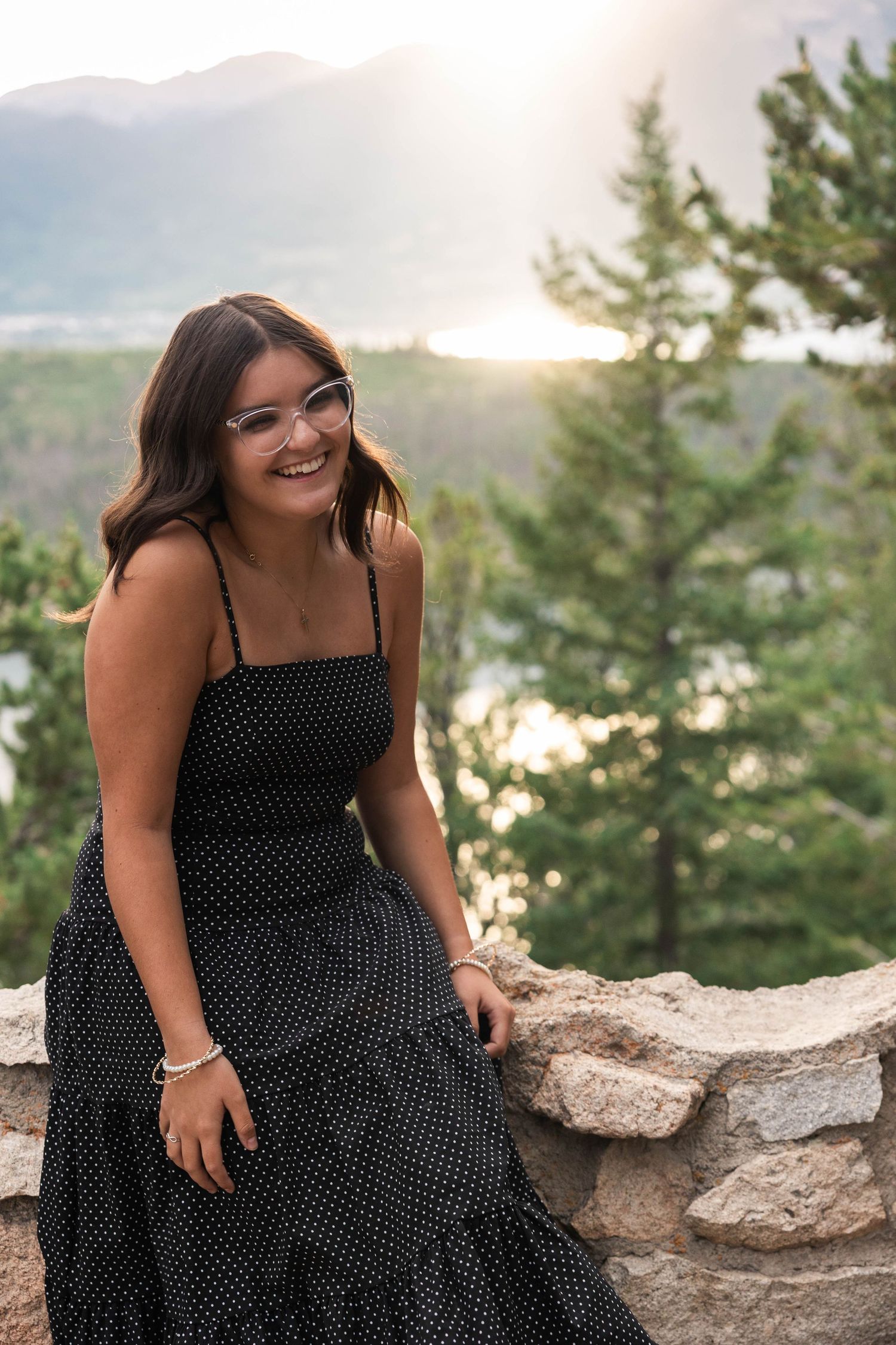 A series of outdoor portrait shots of someone in a black polka dot dress sitting on rocks at sunset by a lake.