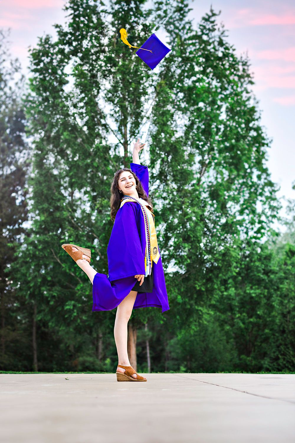 Graduate in purple cap and gown celebrates on an outdoor path during sunset.