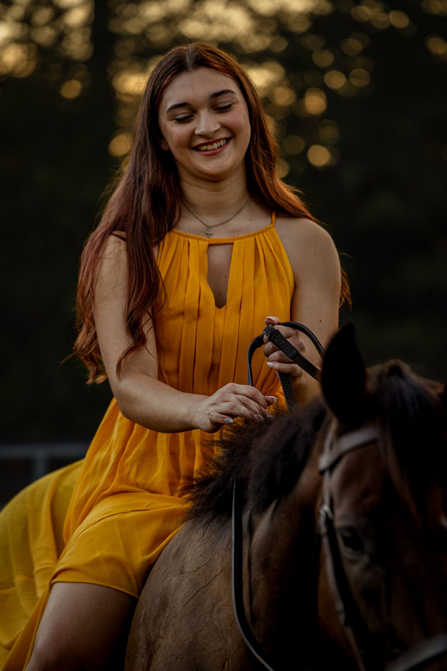 Young woman in yellow dress smiling while riding a brown horse at golden hour.