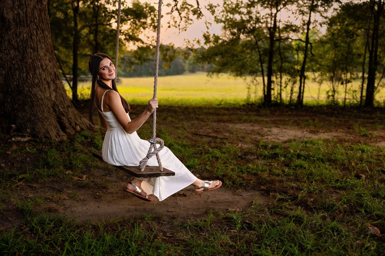 A person in a white dress sits on a forest floor during golden hour as sunlight filters through the trees.