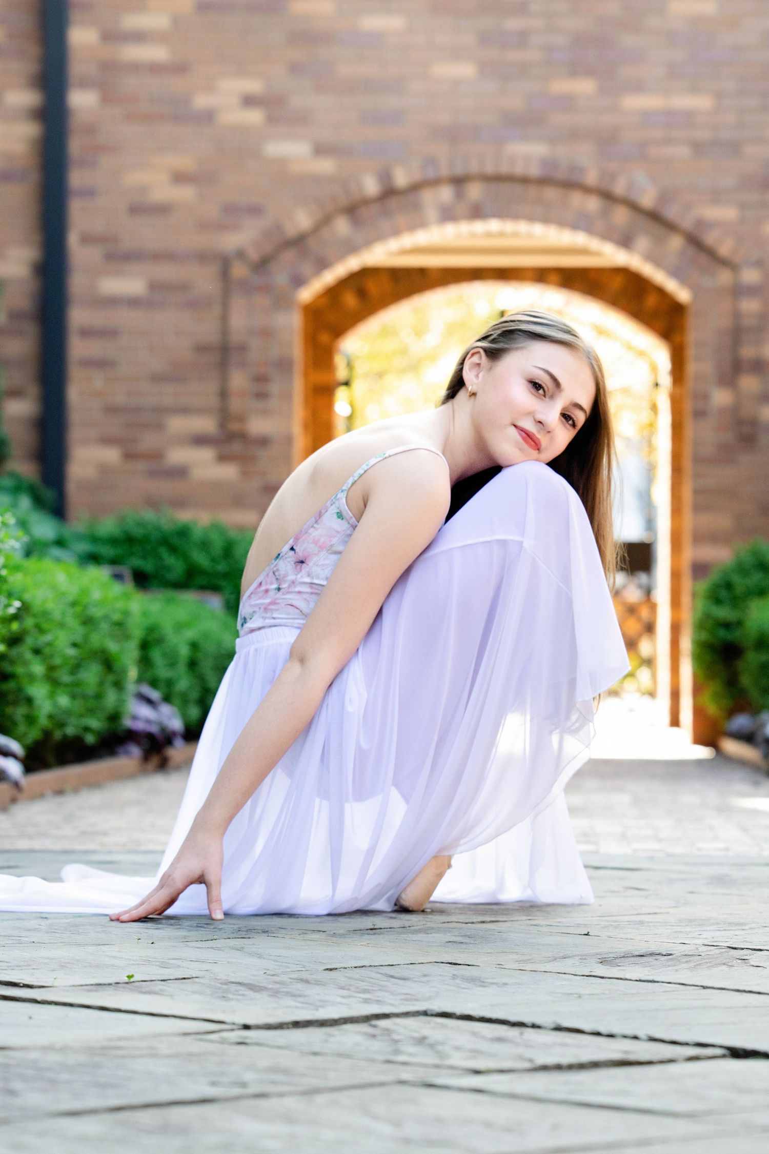 A graceful dancer in a flowing white dress performs an elegant pose on stone pavement near an arched brick wall.