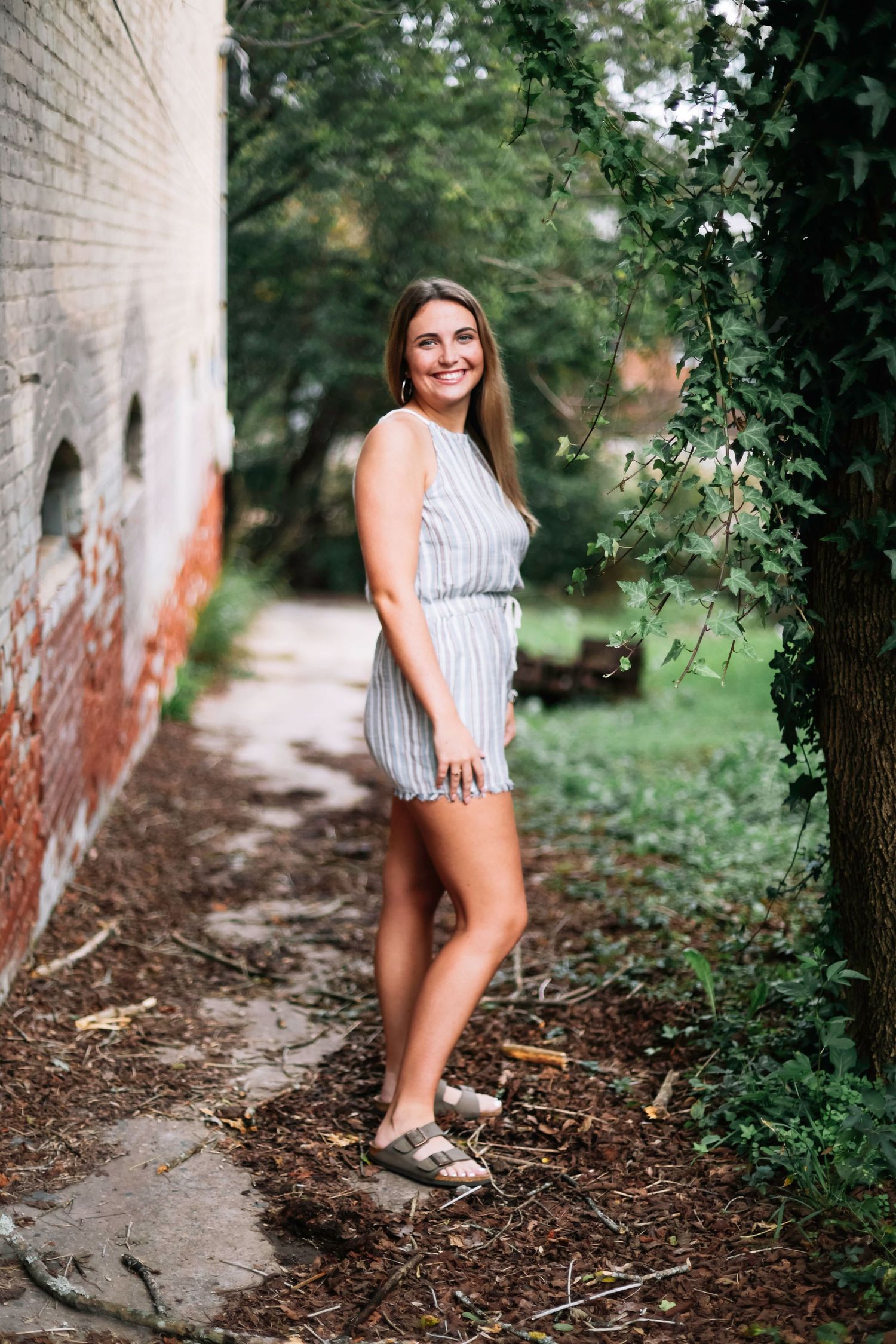 A person in a striped dress poses against a rustic brick wall surrounded by greenery and natural light.