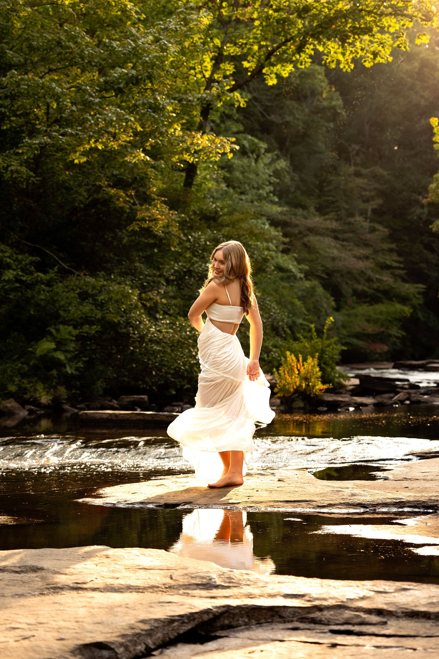 Person in flowing white dress dances on rocky riverbank at sunset.