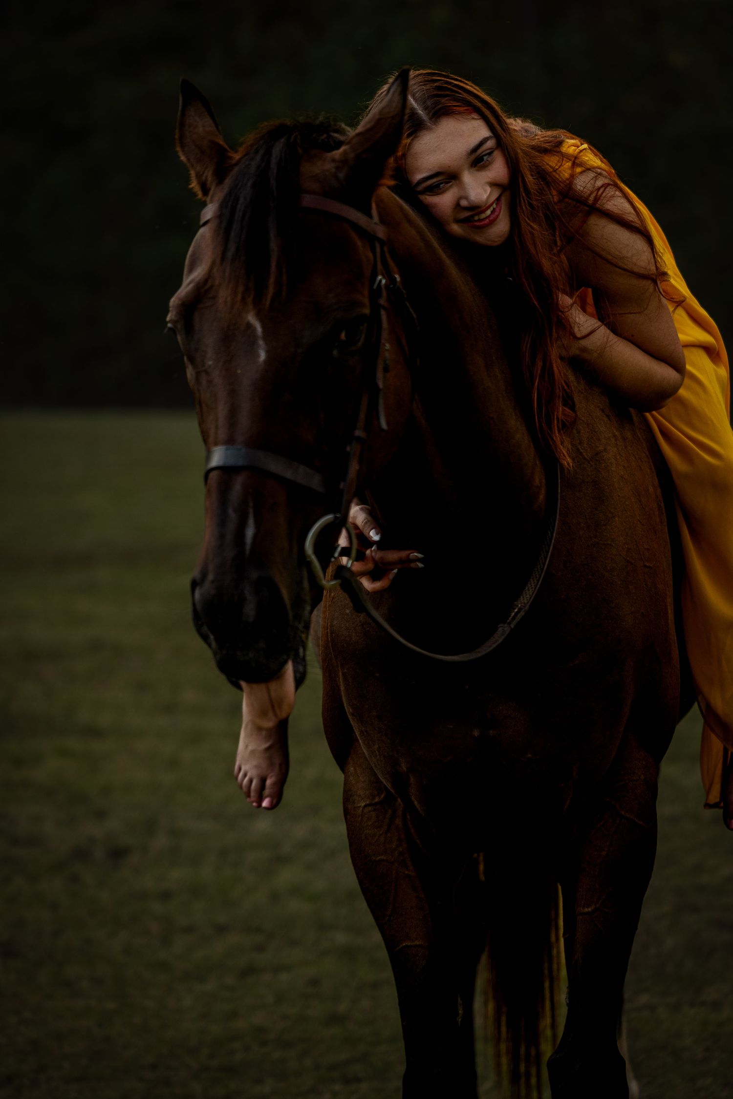 A smiling woman in a golden dress leans affectionately against a brown horse in a dark outdoor field.