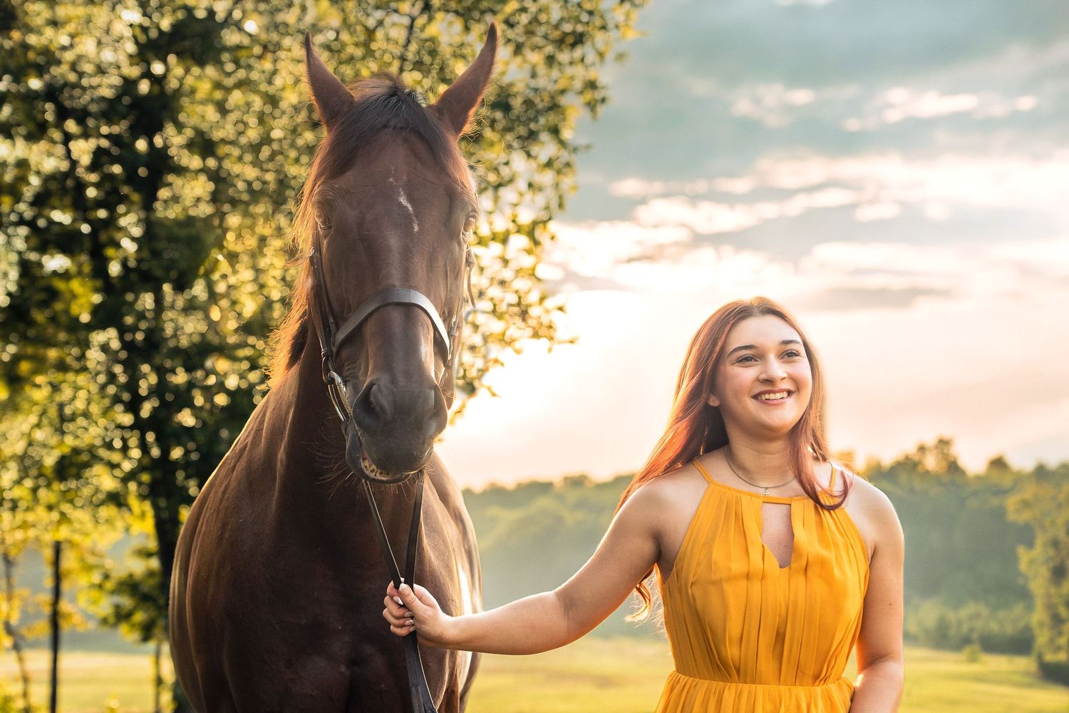 Young woman in yellow dress smiling beside a brown horse outdoors at golden hour.