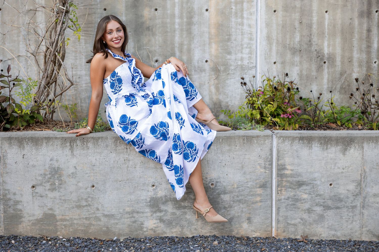 Woman in white and blue floral maxi dress sitting on concrete wall, smiling outdoors.