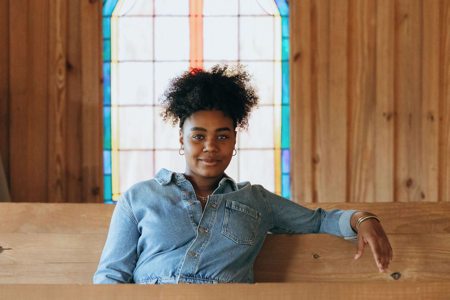 Young Black woman in denim shirt smiling, seated in wooden church pew with colorful stained glass window behind her.