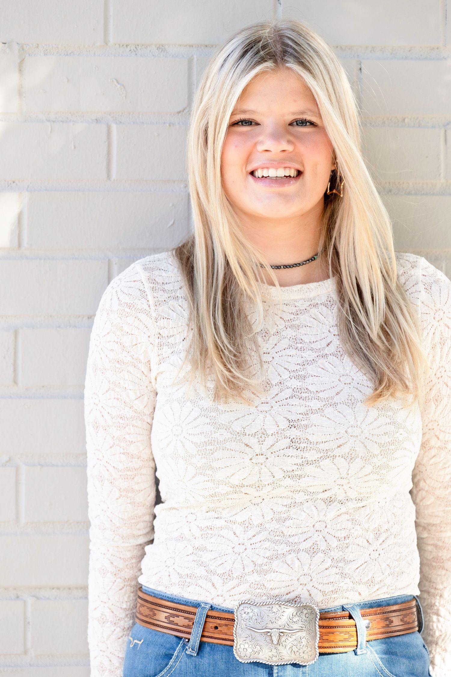 Person wearing white lace top and brown belt poses against white brick wall.