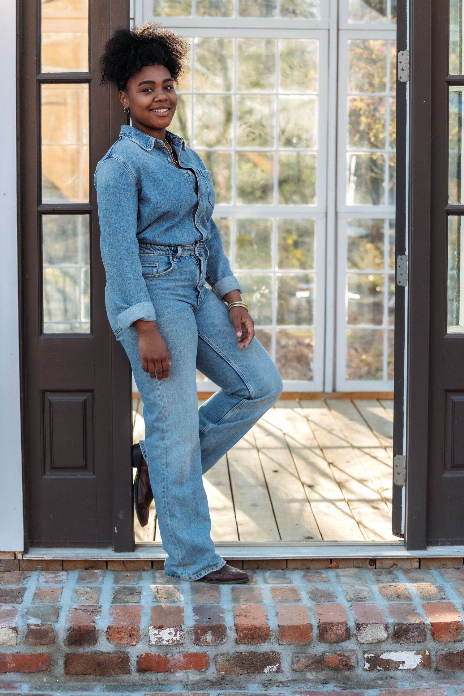 Smiling woman in denim jumpsuit posing by open French doors on brick steps.