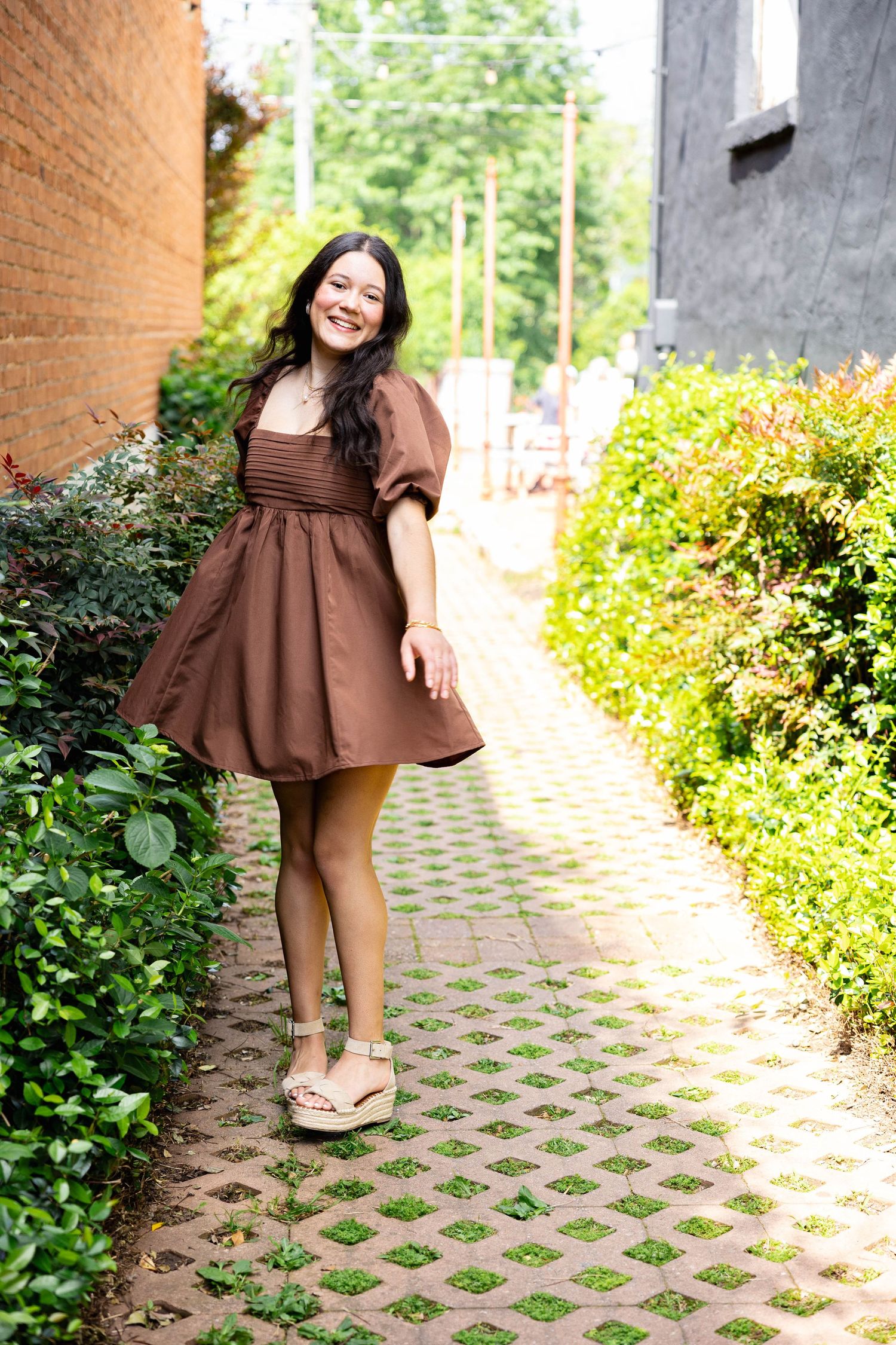 Someone in a brown dress poses cheerfully on a garden path with patterned stone pavers and green landscaping.