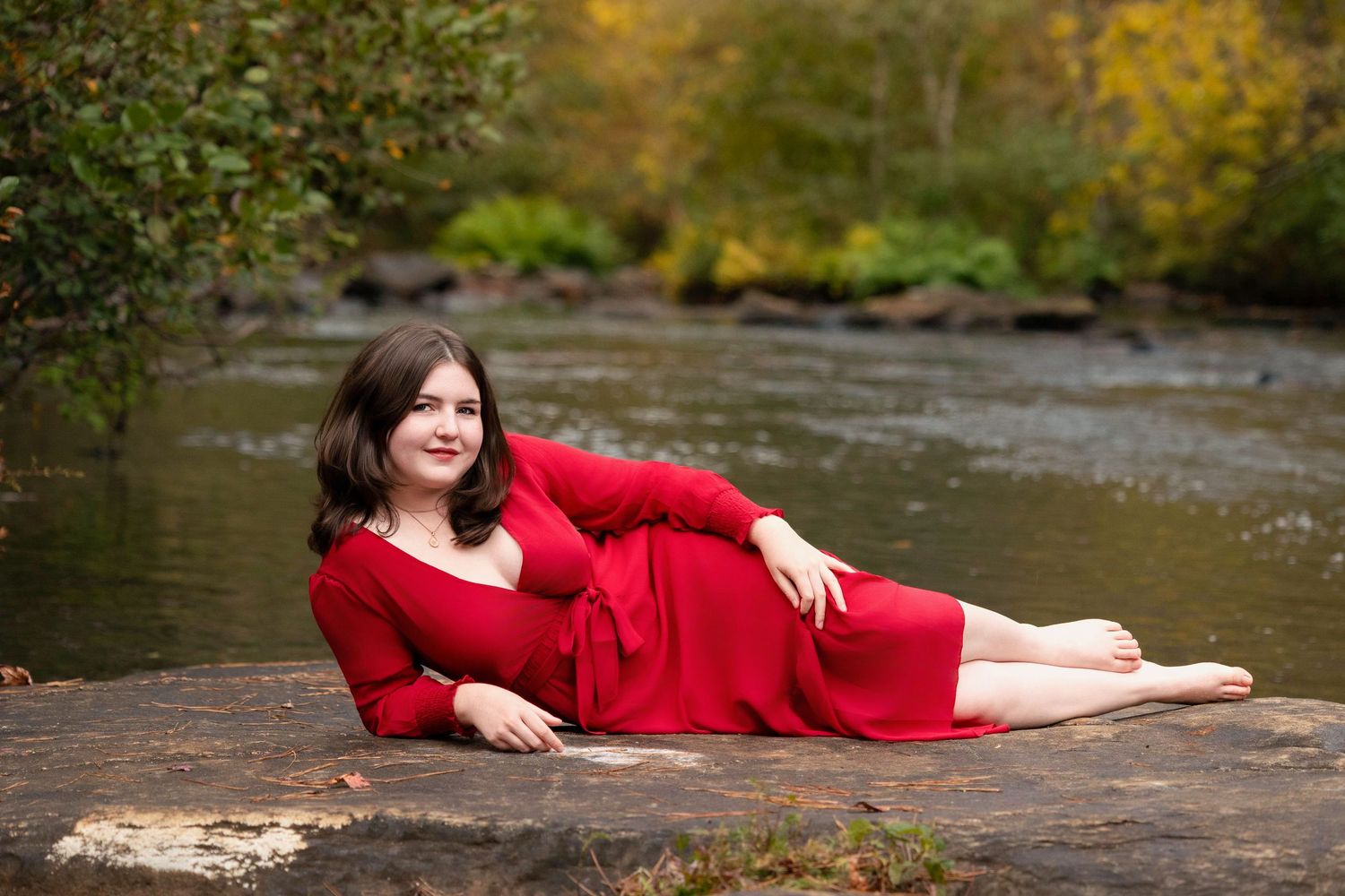 A figure in a flowing red dress poses gracefully on a stone surface near a rushing stream in autumn.