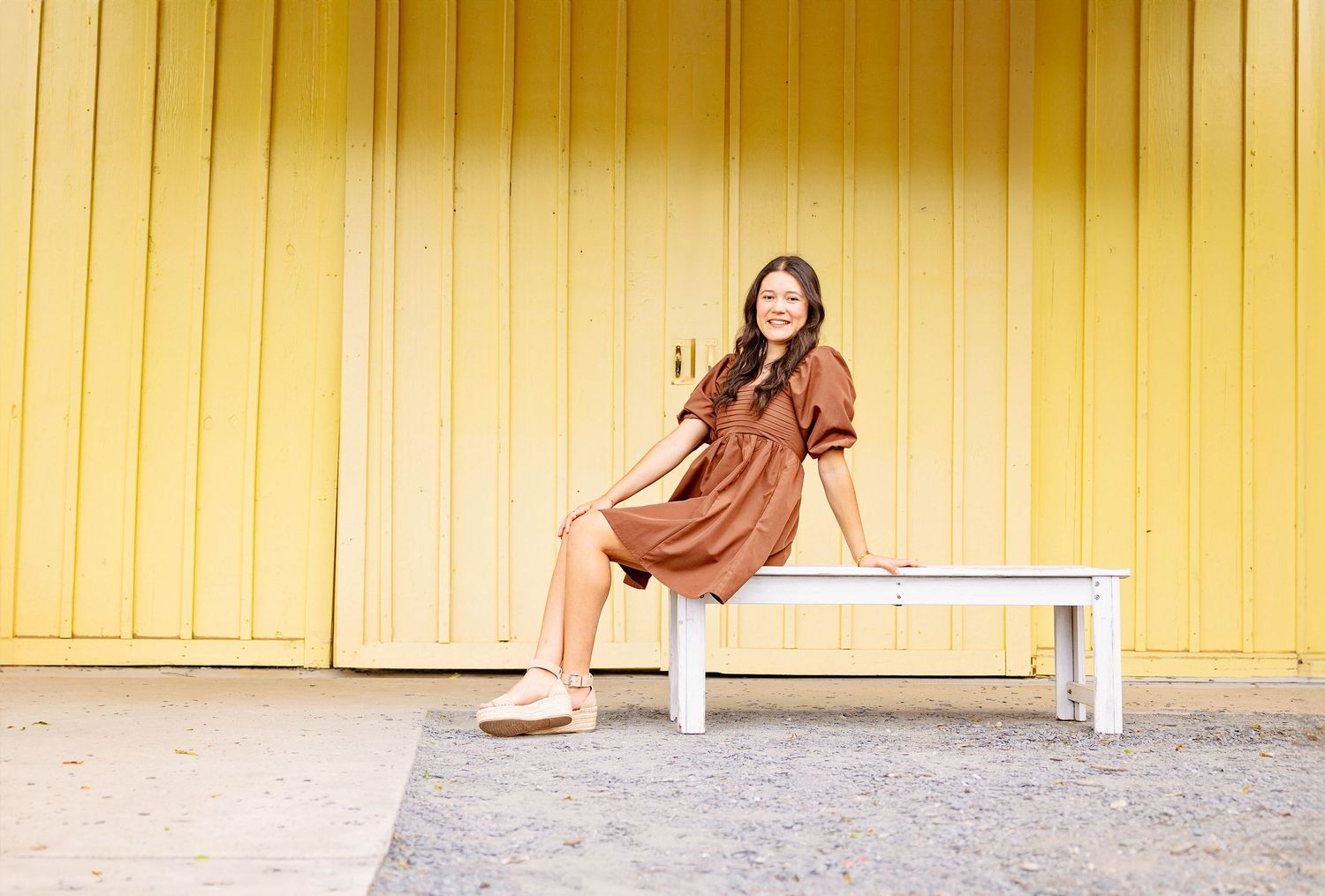 Woman in casual business attire poses confidently against a yellow wall backdrop in natural light.