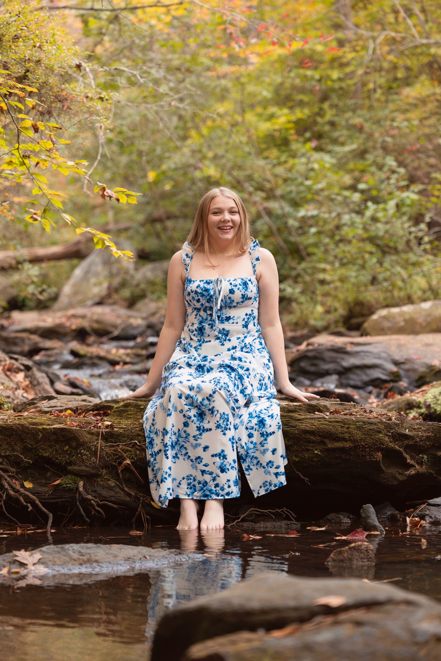 A woman in a blue floral dress sits on a fallen log by a stream surrounded by autumn foliage.