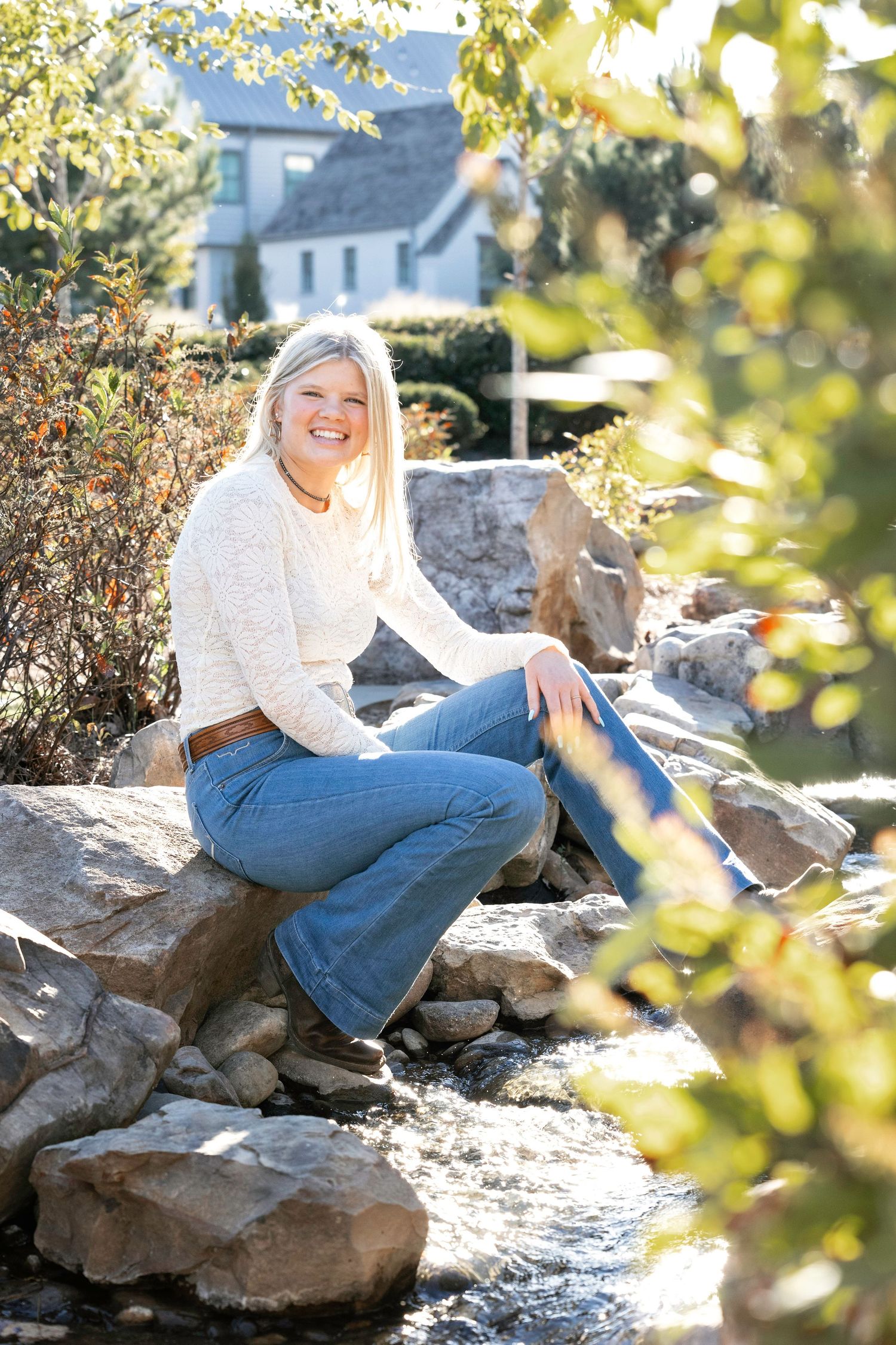 Person in white sweater and jeans sits casually on large rocks in outdoor setting with sunlit foliage.