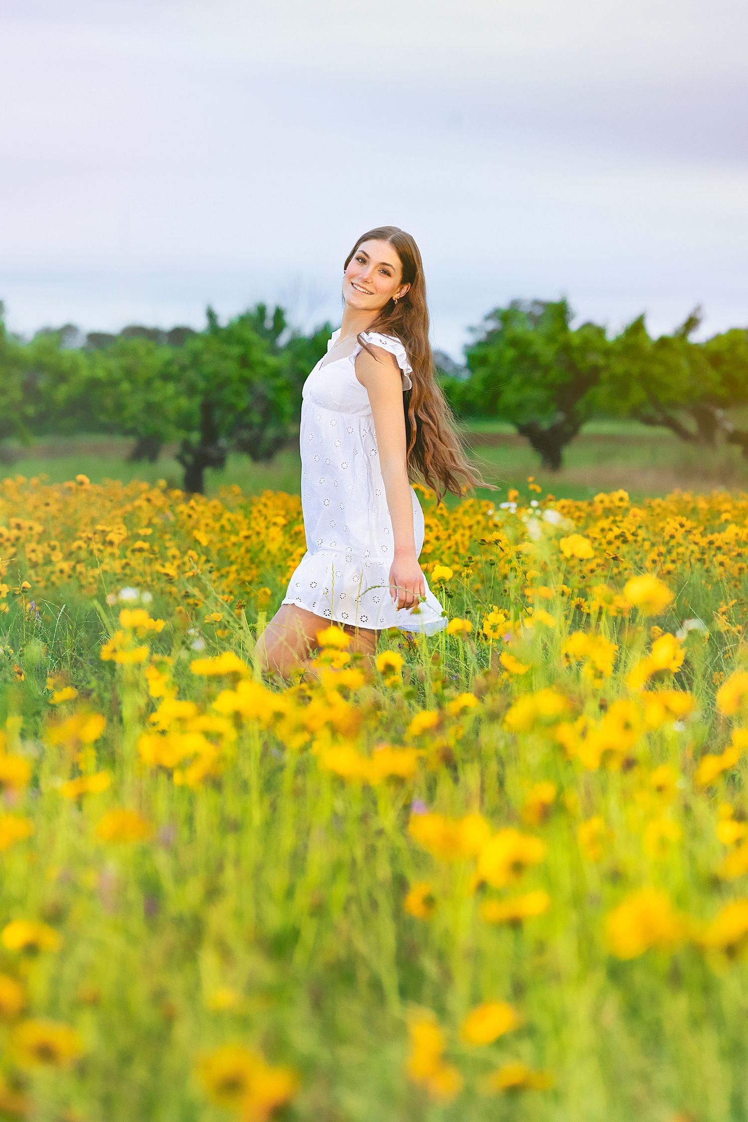 Young woman in white dress smiling in a vibrant yellow wildflower field with green trees in background.