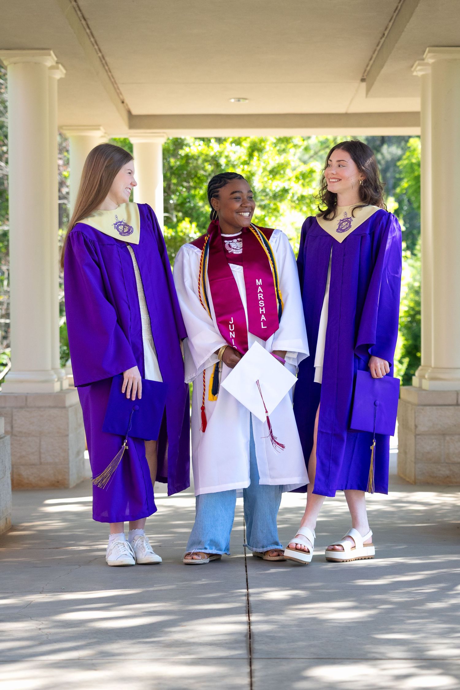 Three graduates in purple and white gowns smile together under a columned walkway at an outdoor graduation ceremony.