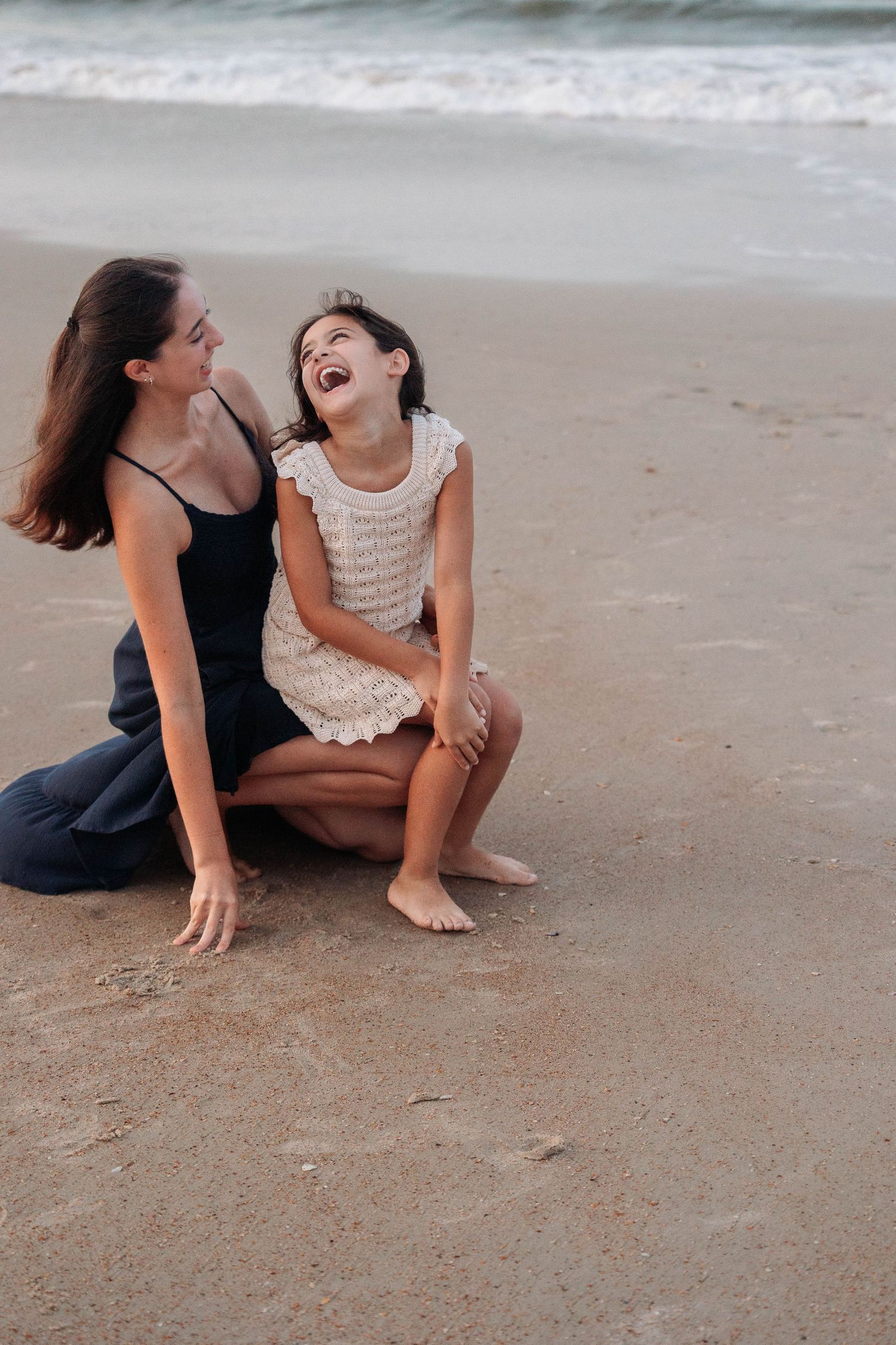 Two people in casual attire share a joyful moment while sitting on the sandy beach at sunset.