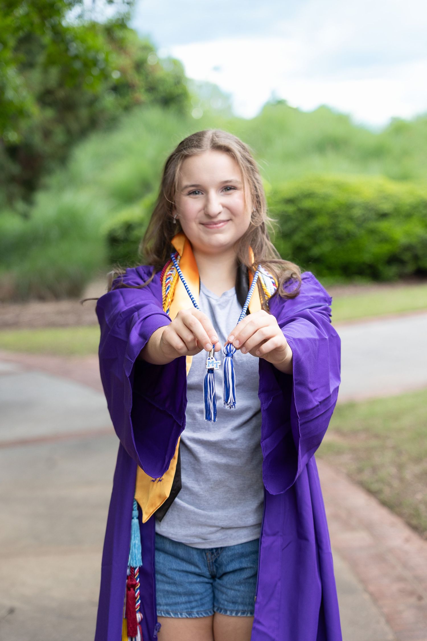 A person in a purple graduation gown and gold stole smiles while holding their graduation cords outdoors.